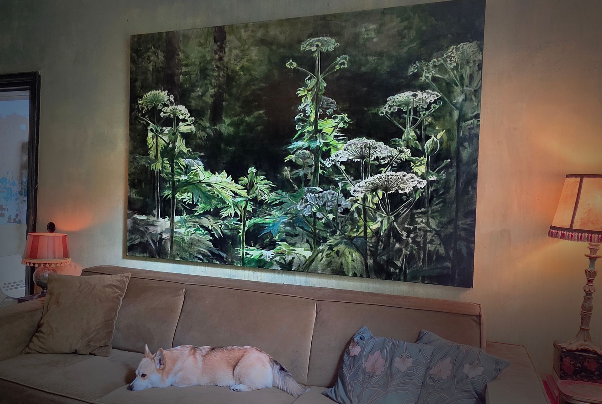 Woonkamer met een groot ingelijst boslandschap boven een beige bank en twee brandende tafellampen.