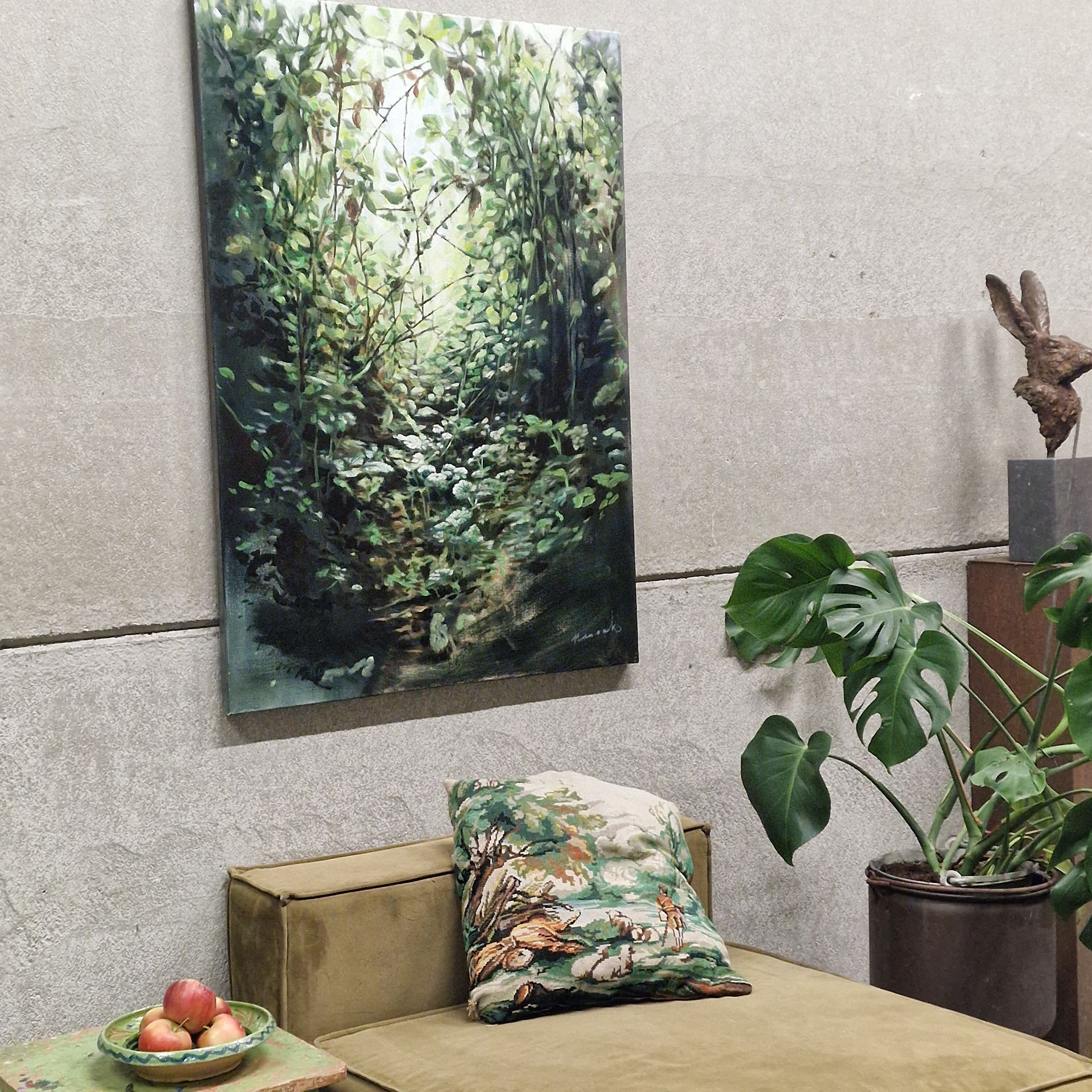 Moderne loungehoek met een schilderij van een bos, een monstera in een pot, een kussen en een klein fruitschaaltje.