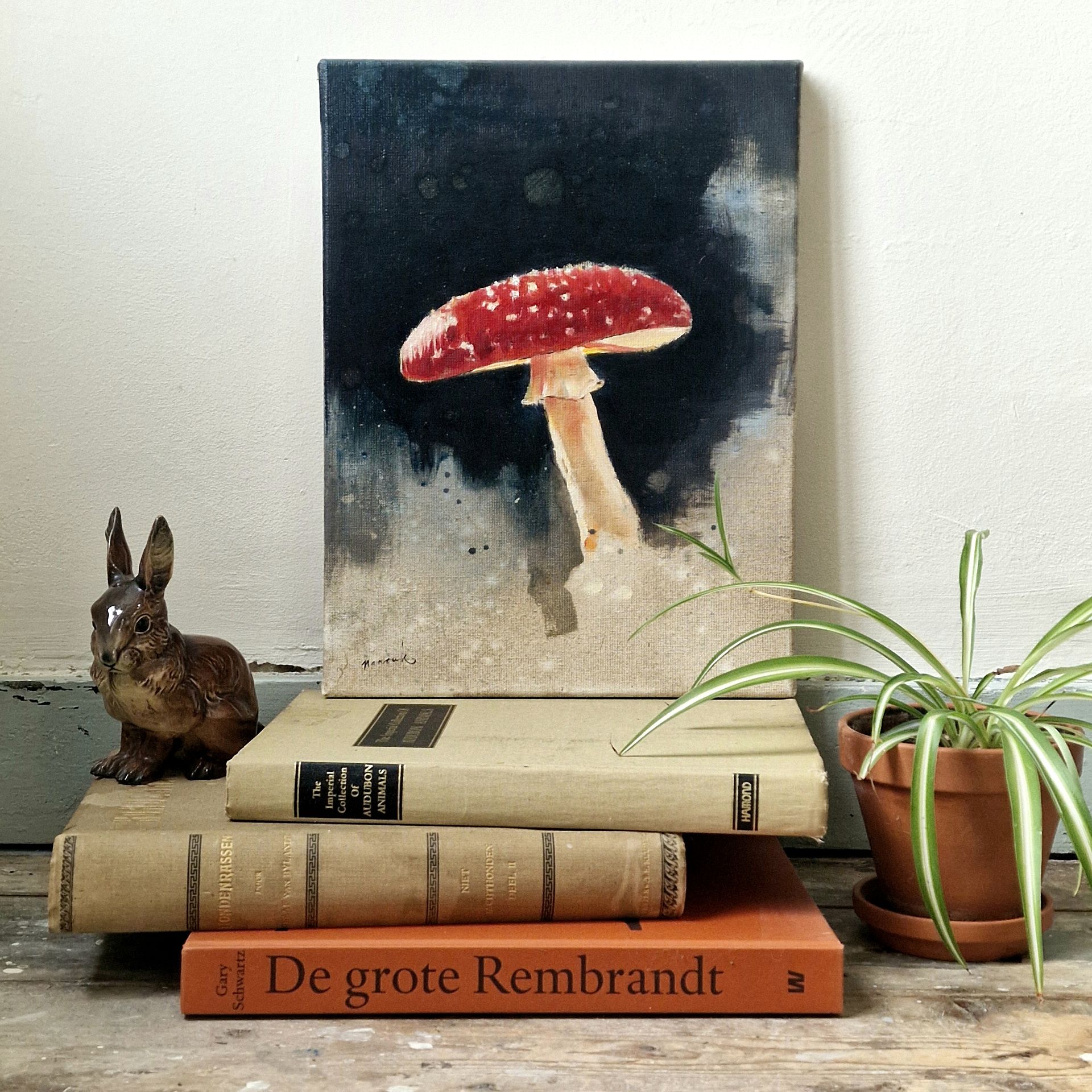 Een stilleven met paddenstoelenkunst, boeken, een konijnenbeeldje en een potplant op een houten ondergrond.