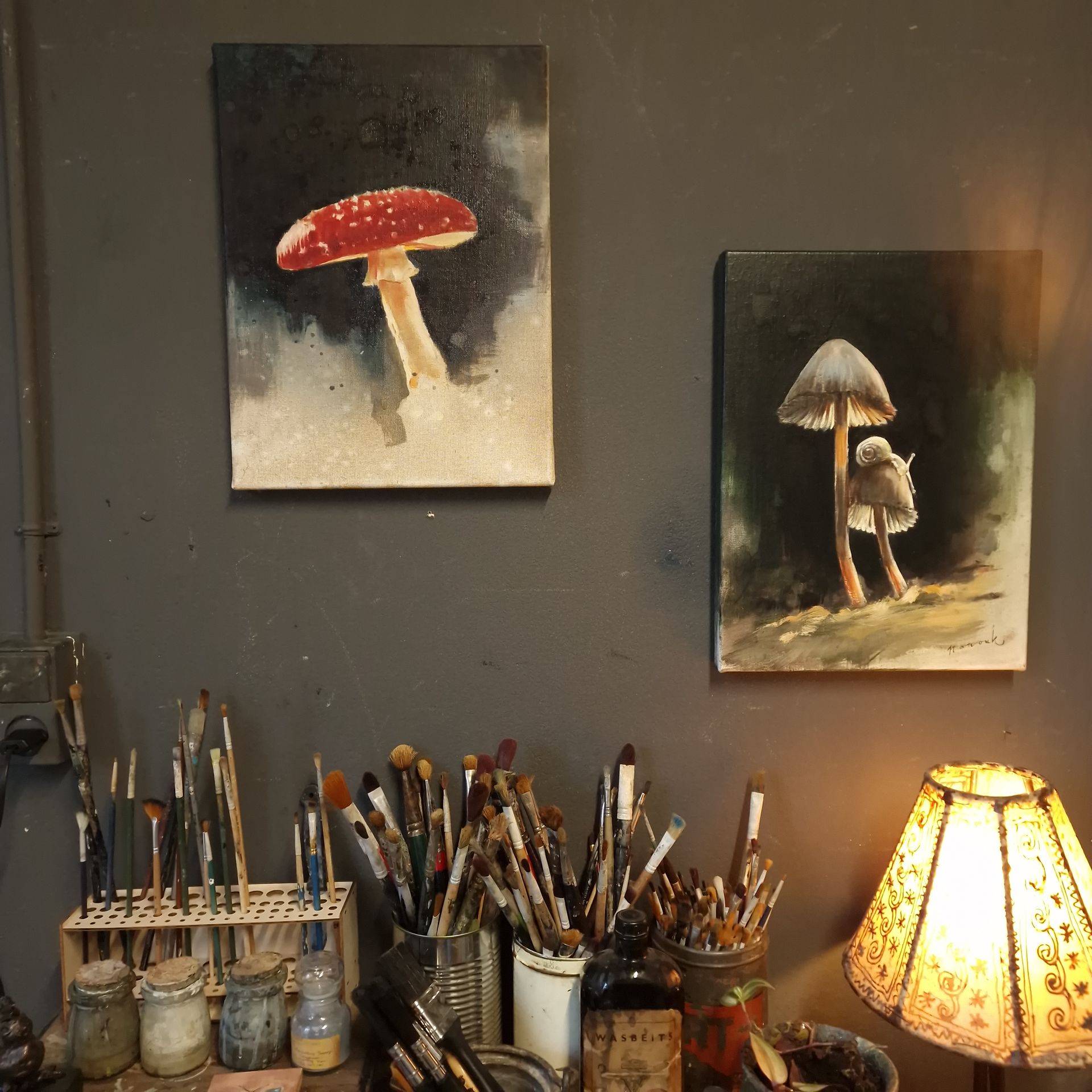 Muur van een kunststudio met twee schilderijen van paddenstoelen boven een tafel vol penselen en een gloeiende lamp.