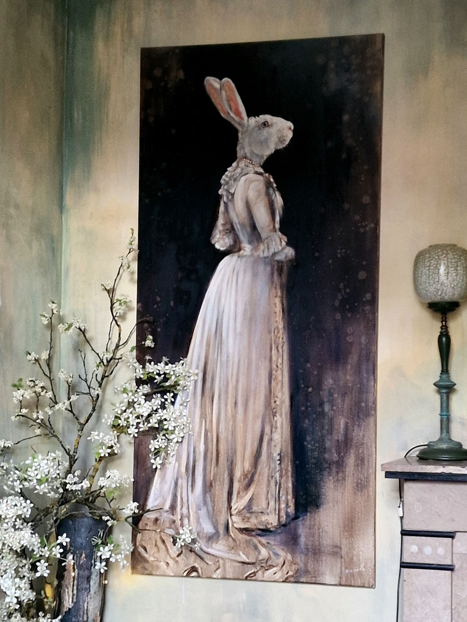 Schilderij van een konijn in een lange jurk op een donker doek, naast witte bloesems en een lamp.
