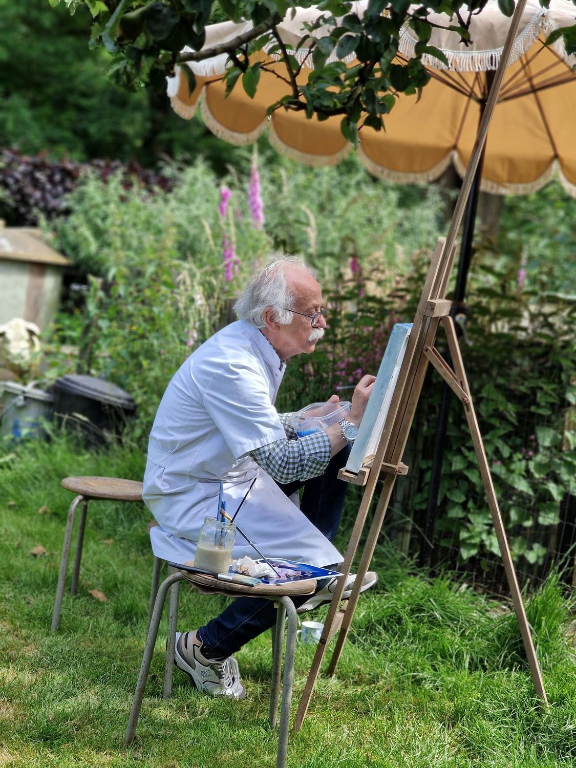 Een man zit op een krukje en schildert op een ezel in een tuin.