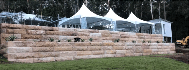 Stone retaining wall with white tents and a small building in a grassy area. Trees in the background.