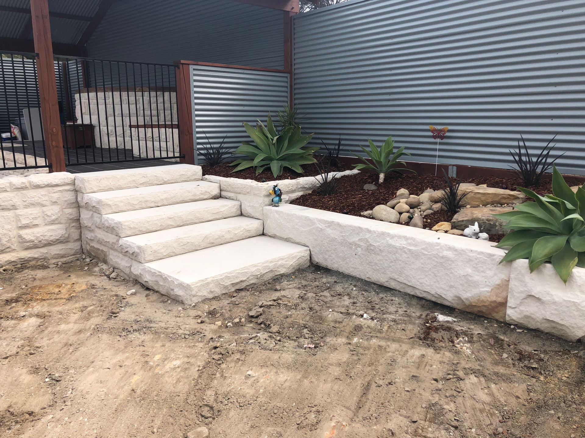 Stone steps lead to a raised garden bed with plants, mulch, and a metal corrugated wall.