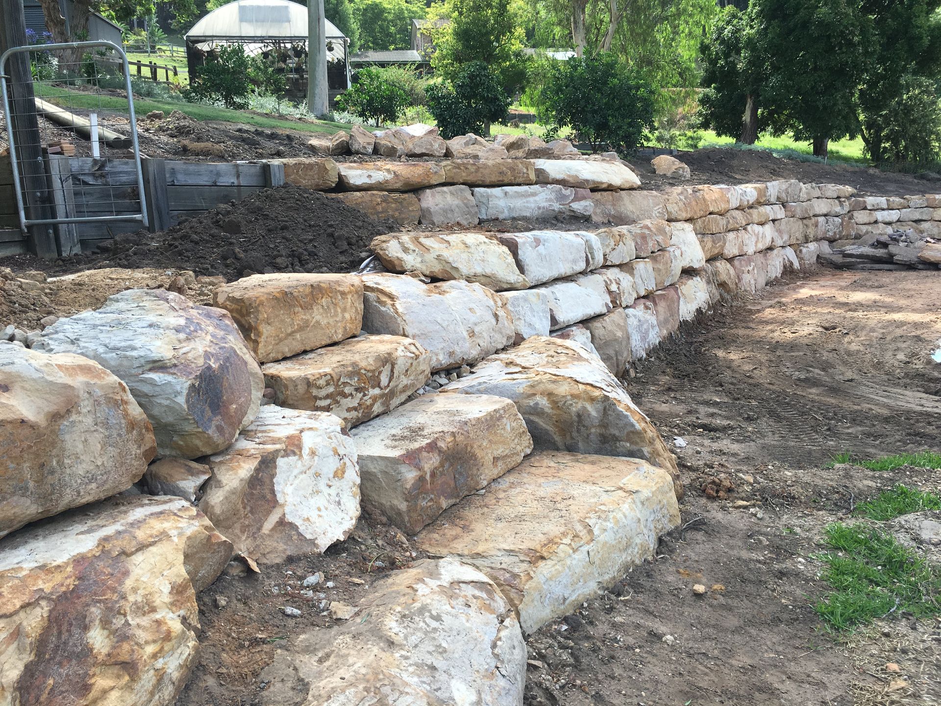 Stone retaining wall under construction on a hillside with dirt and surrounding landscaping.