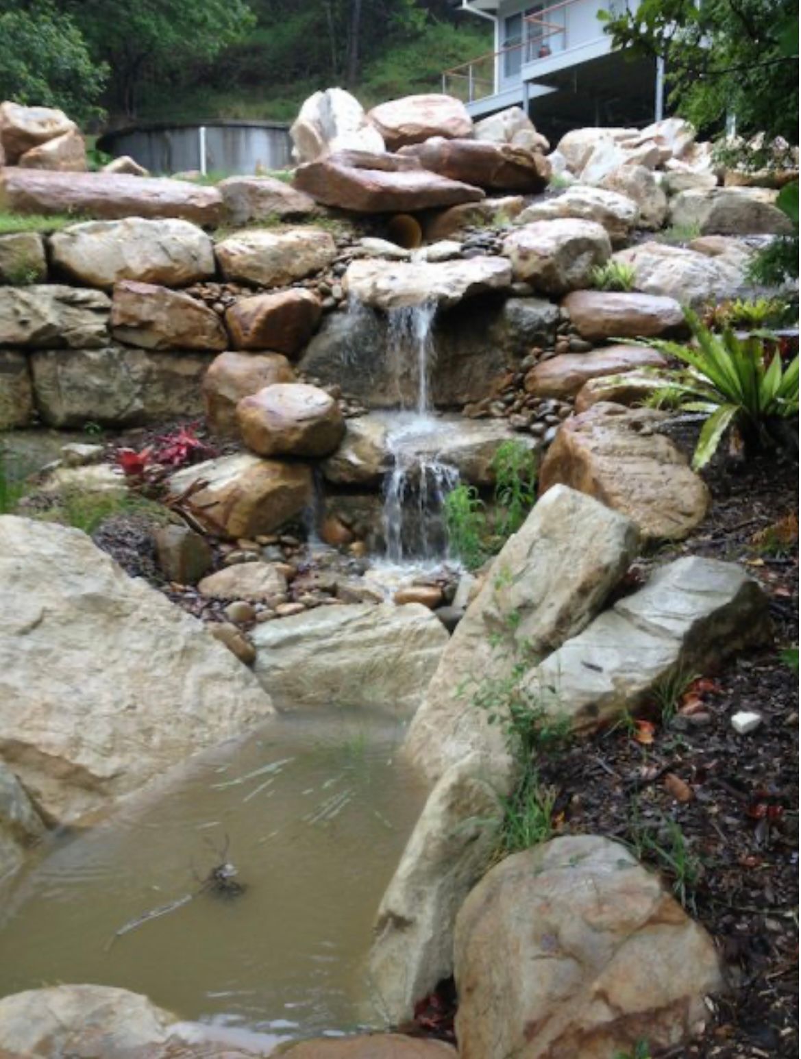 Waterfall cascading over large rocks into a murky pond, with plants and a house visible.