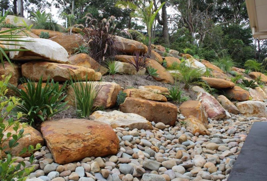 Rockery landscaping with large sandstone boulders, small pebbles, and native plants on a hillside.