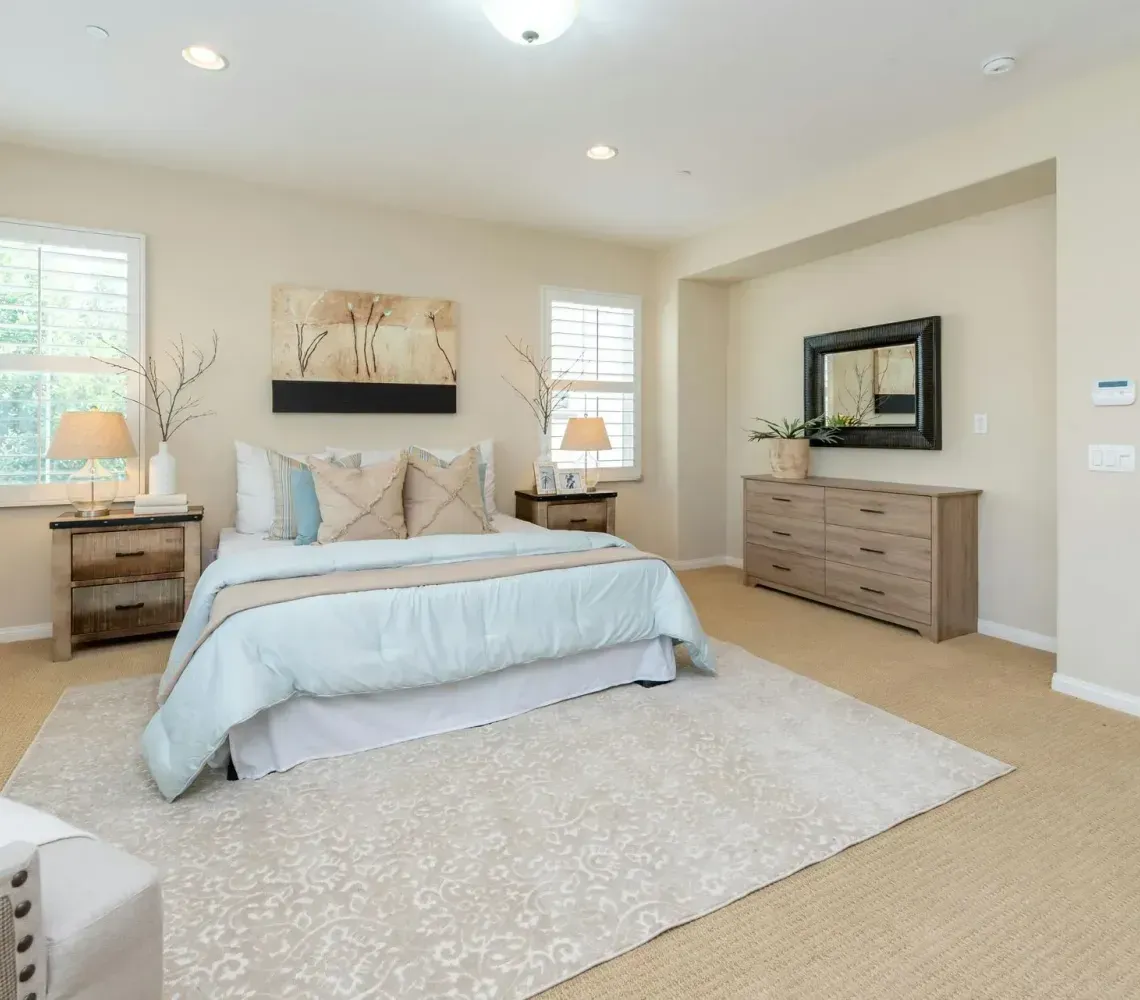 A bright bedroom featuring a queen bed with a light blue comforter, two wooden nightstands, and a matching dresser.