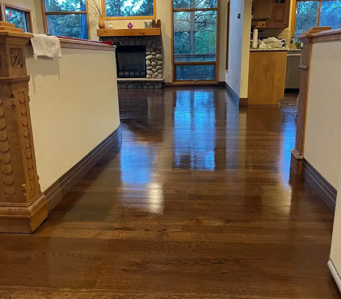 A sunlit room with high ceilings, polished dark hardwood floors, a stone fireplace, and views of a forested exterior.
