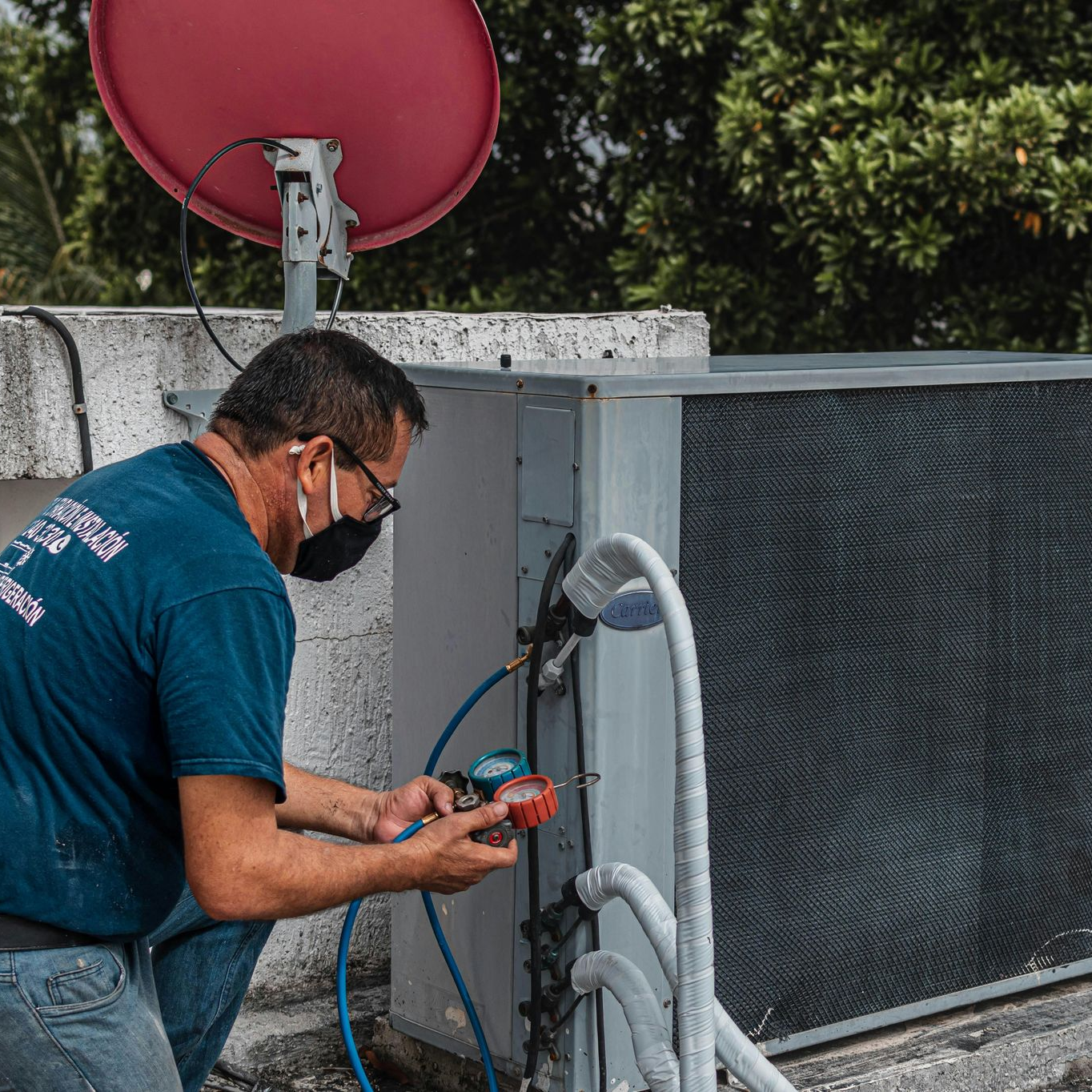 A technician wearing a face mask services an outdoor HVAC unit with a manifold gauge set on a rooftop.