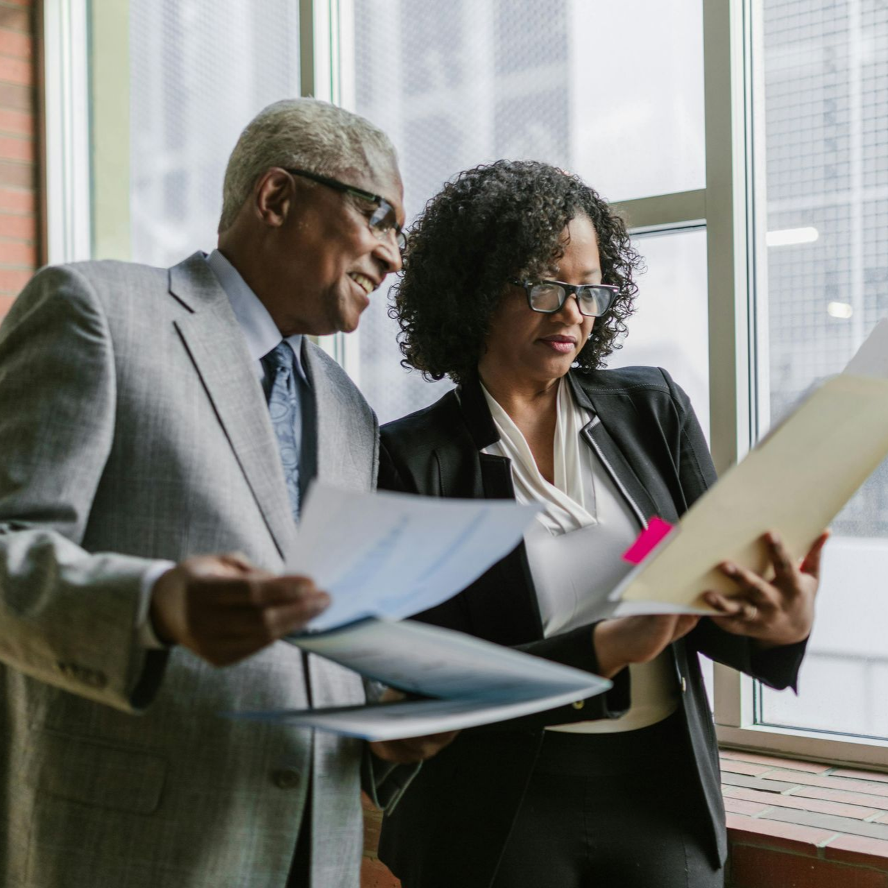 Two colleagues in business attire review documents together in a brightly lit office near a window.