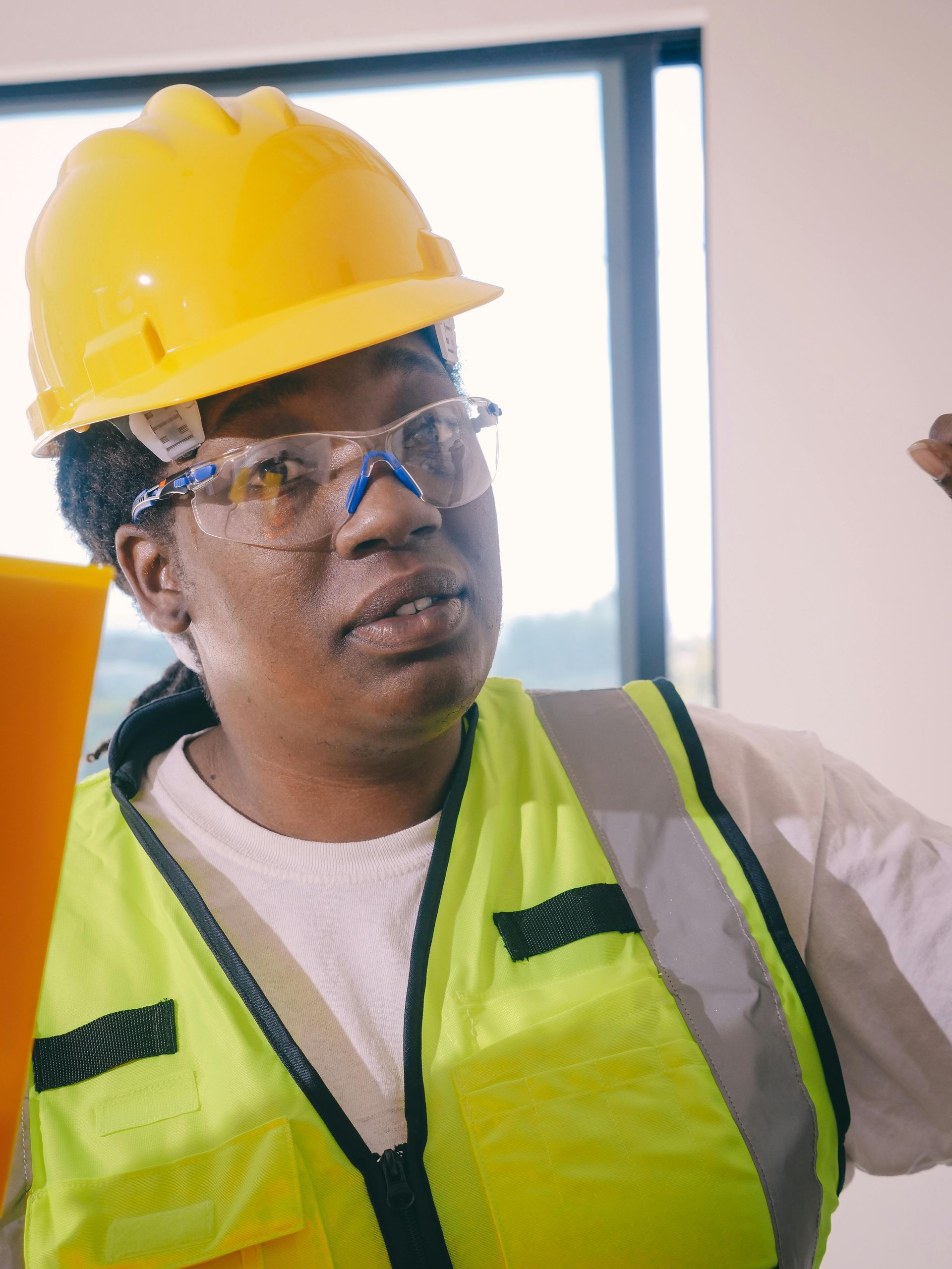 A person wearing a bright yellow hard hat, safety glasses, and a neon yellow safety vest, looking up in an interior room.