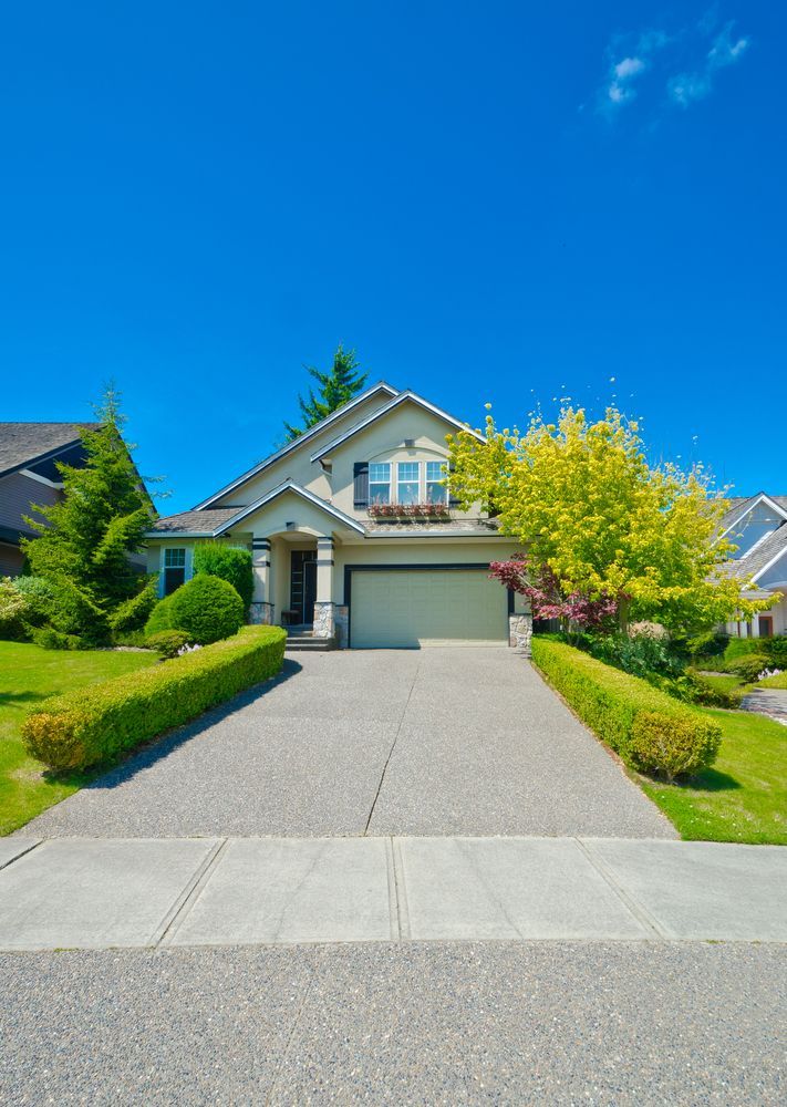 Suburban house with a two-car garage, manicured hedges, and a wide concrete driveway under a bright blue sky