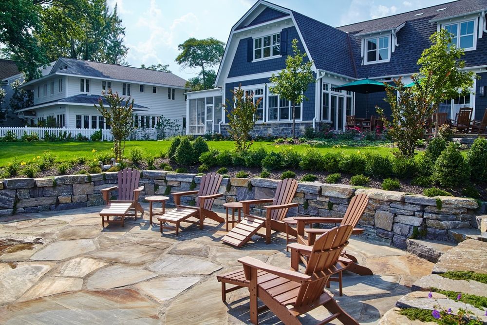 Stone patio with wooden chairs in a landscaped yard, with two blue-and-white houses in the background