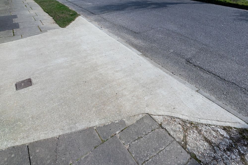 Cracked sidewalk corner meeting a sloped asphalt road and curb.