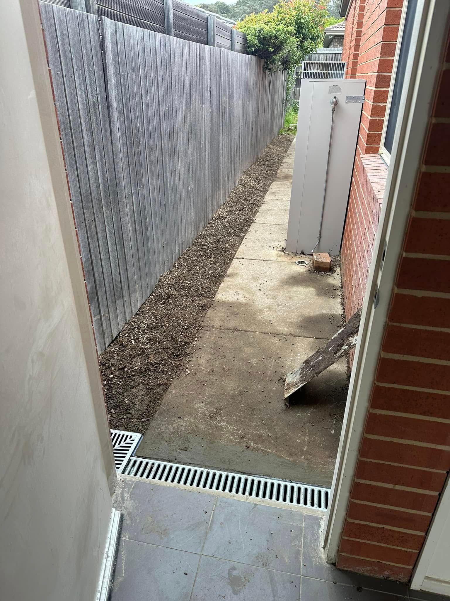 Narrow pathway alongside a wooden fence and brick wall, with leaves scattered on the ground and a drainage grate in the foreground — B3 Plumbing in Flynn, ACT
