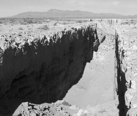 A deep rocky canyon splitting a barren desert plain beneath distant mountains.