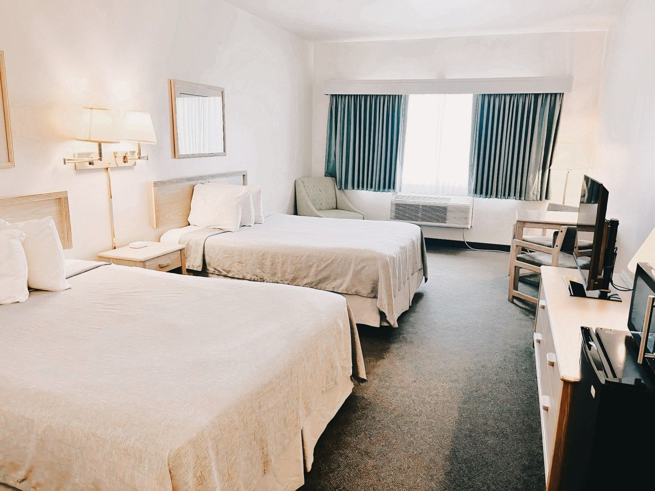 Bright hotel room with two beds, sofa, TV, dresser, and balcony doors with curtains.