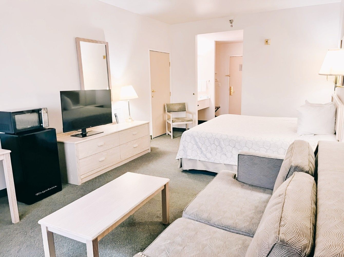 Bright hotel room with two beds, sofa, TV, dresser, and balcony doors with curtains.