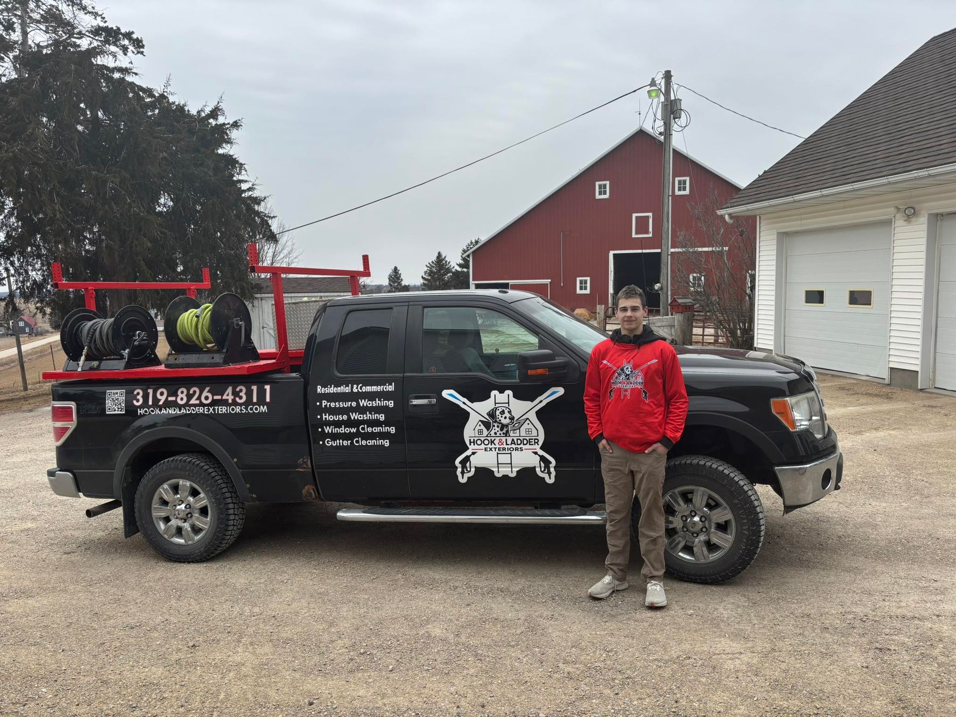 Man standing beside a black pickup truck with red ladder racks in a rural driveway