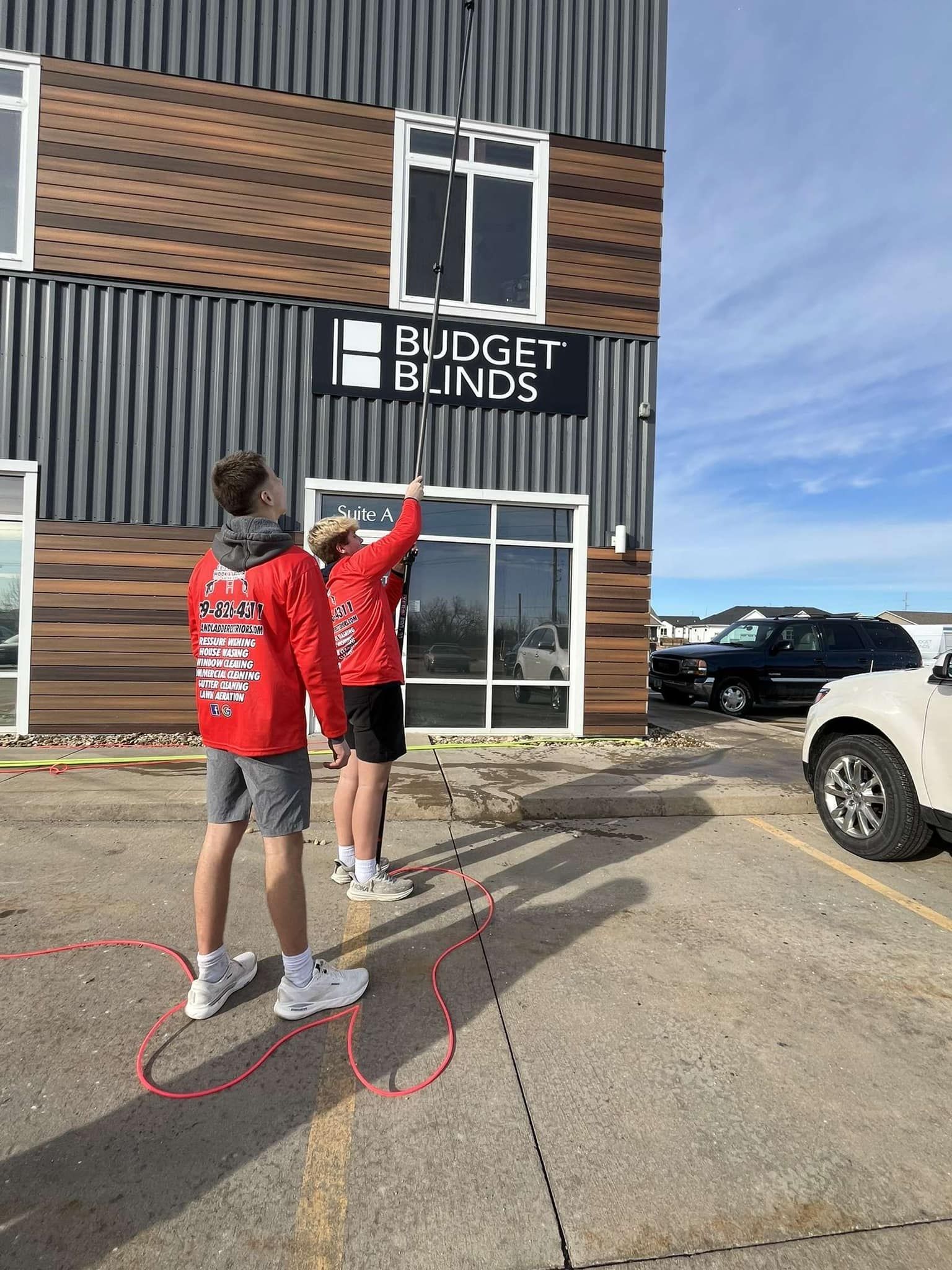Two people in red shirts stand outside a Budget Blinds storefront holding up window blinds.