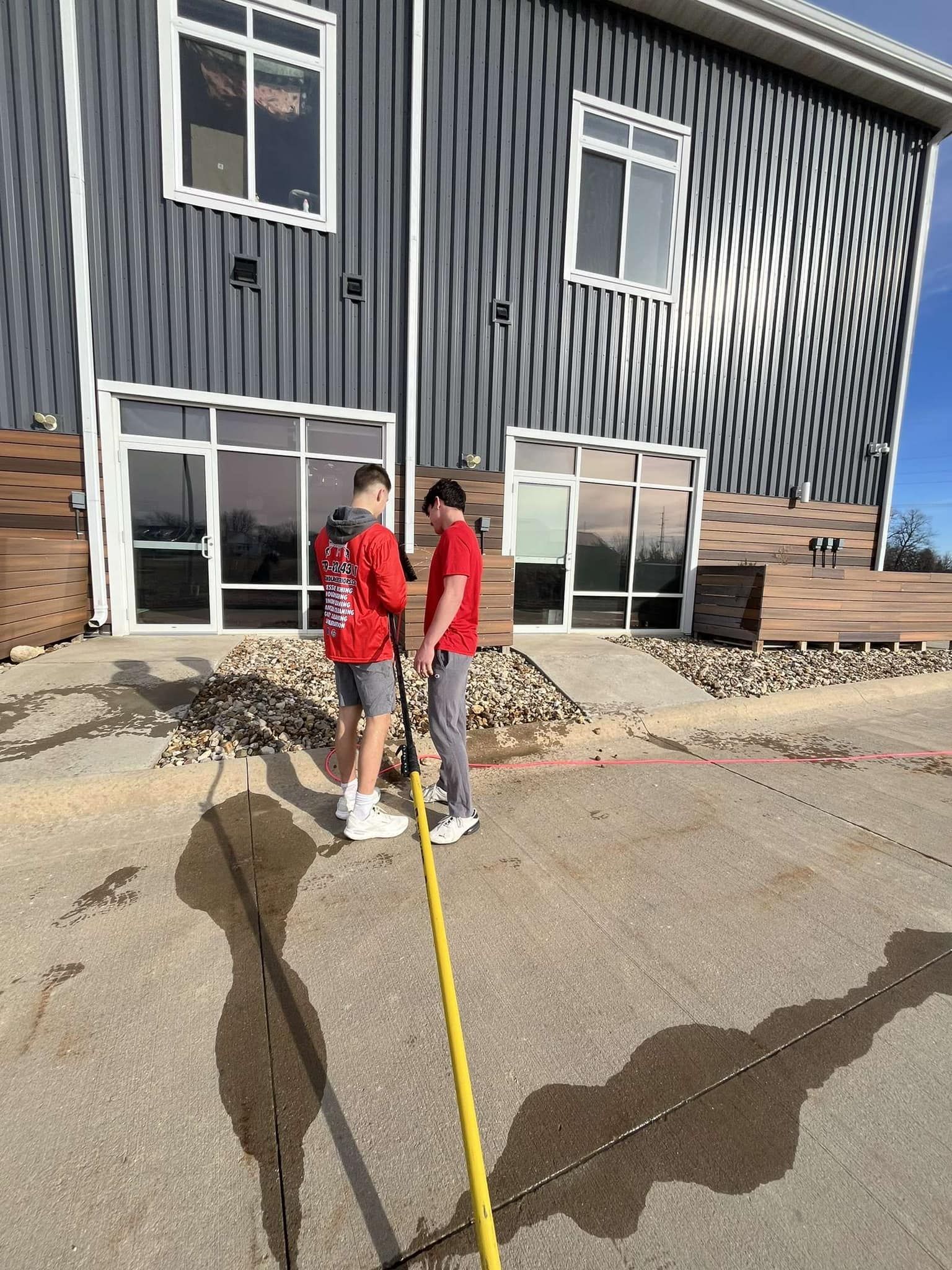 Two people in red jackets stand on a paved area beside a gray building with a yellow hose or rope between them.