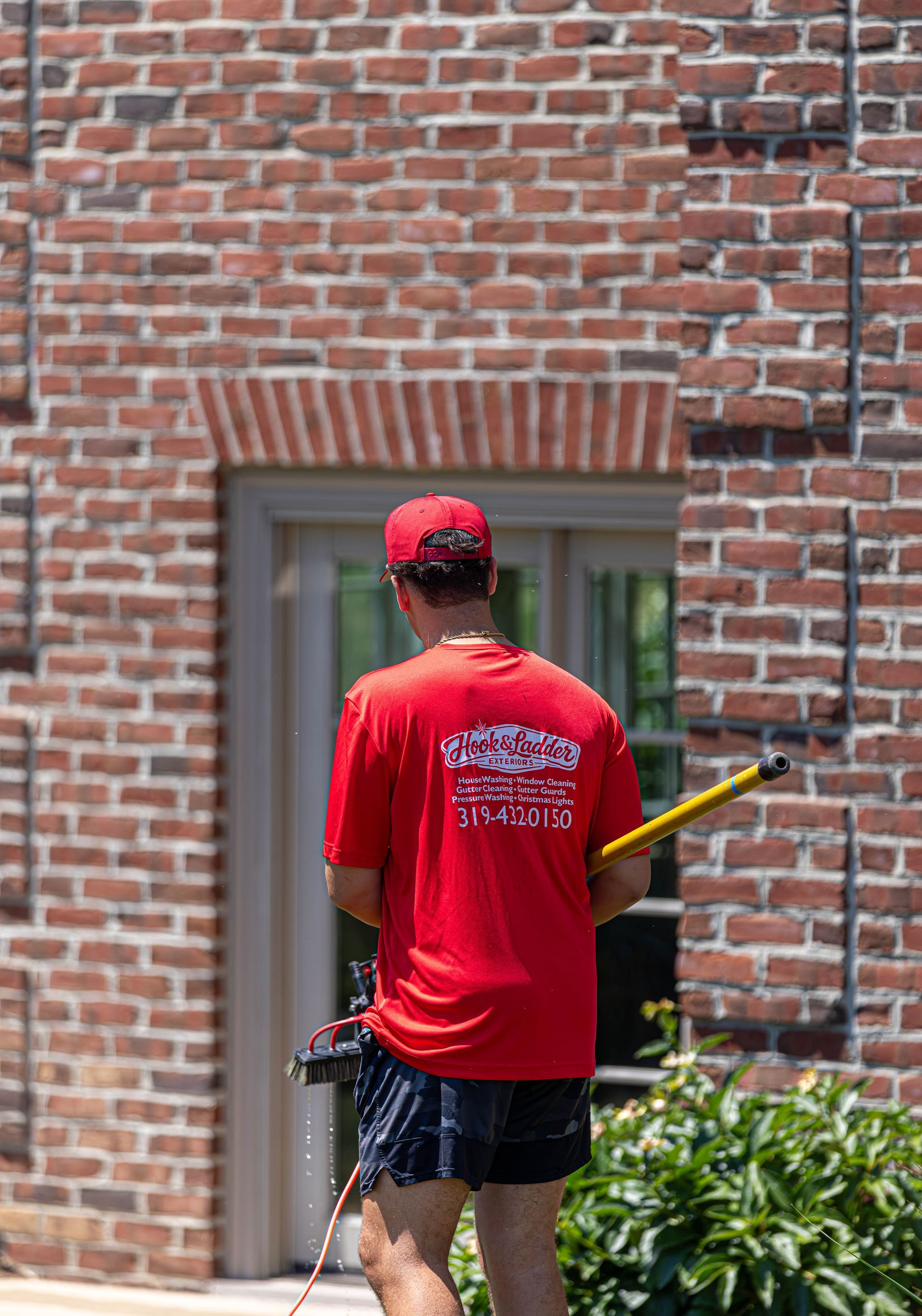 Person in red shirt and cap using a weed trimmer beside a brick building.