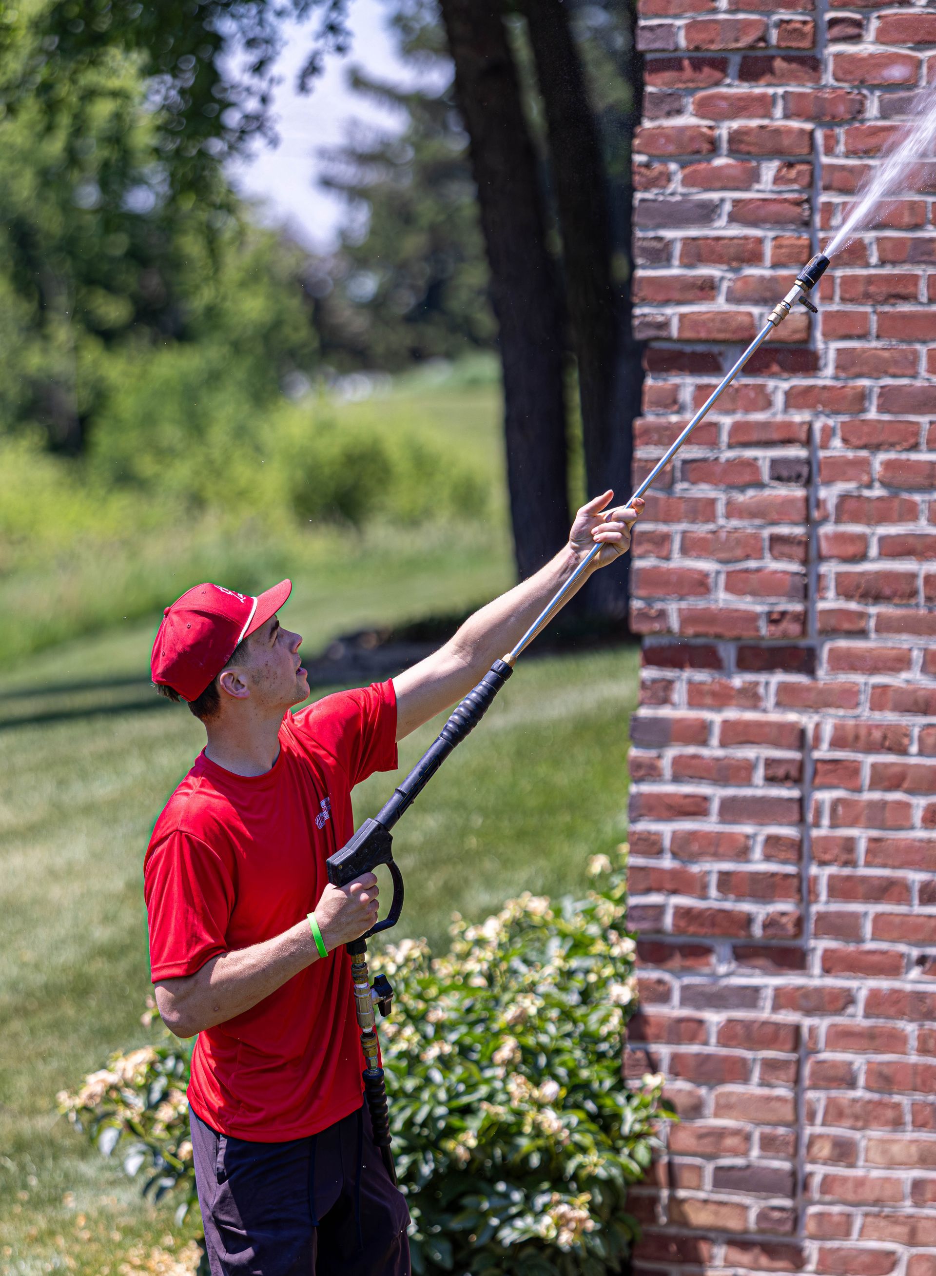 Person in a red shirt using a long pole to clean a brick wall outdoors