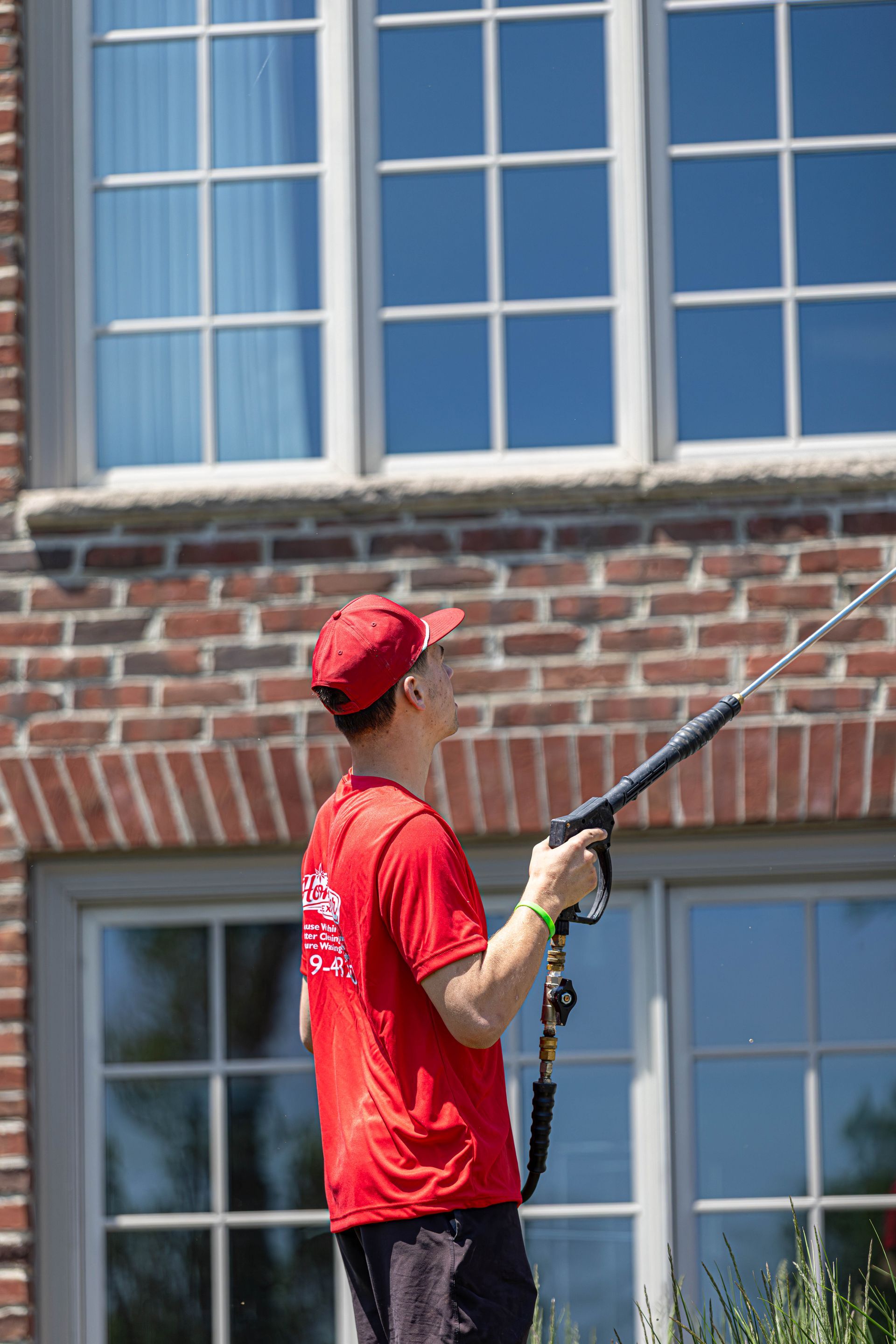 Person in red shirt and cap using a long pole to clean or inspect a brick building’s windows