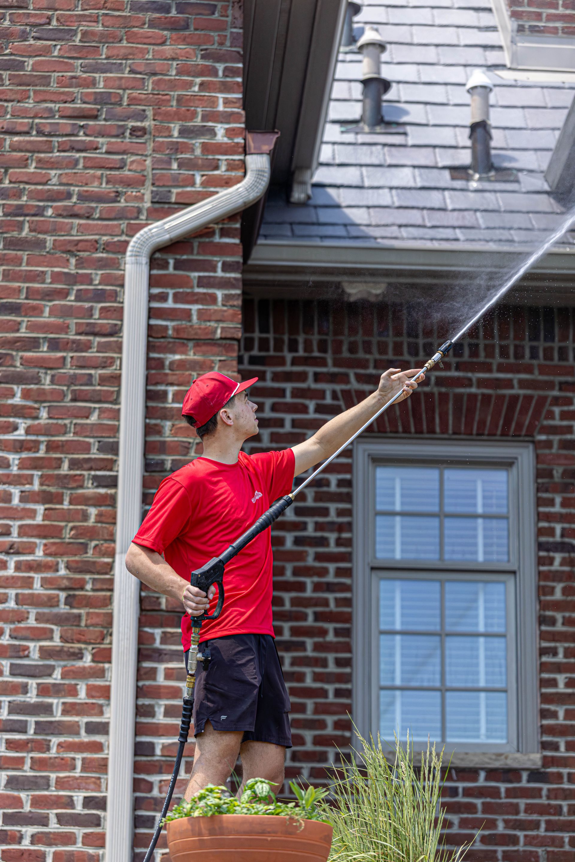 Person in red shirt and hat spray-washes a brick house wall near a window.