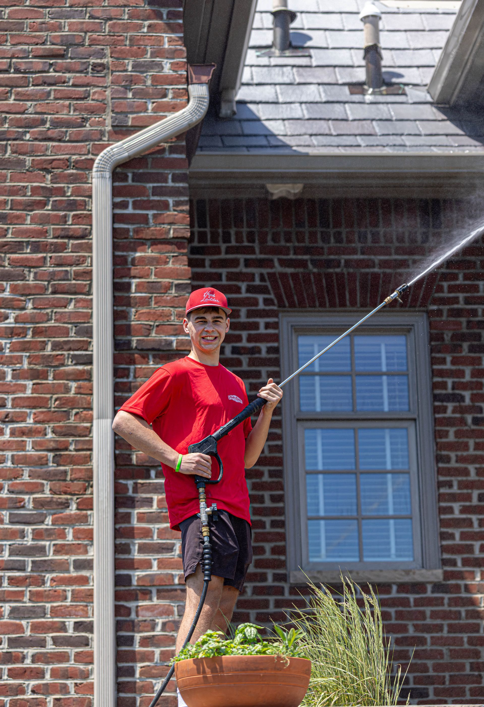 Person in red shirt and cap washing a brick house window with a pressure washer.
