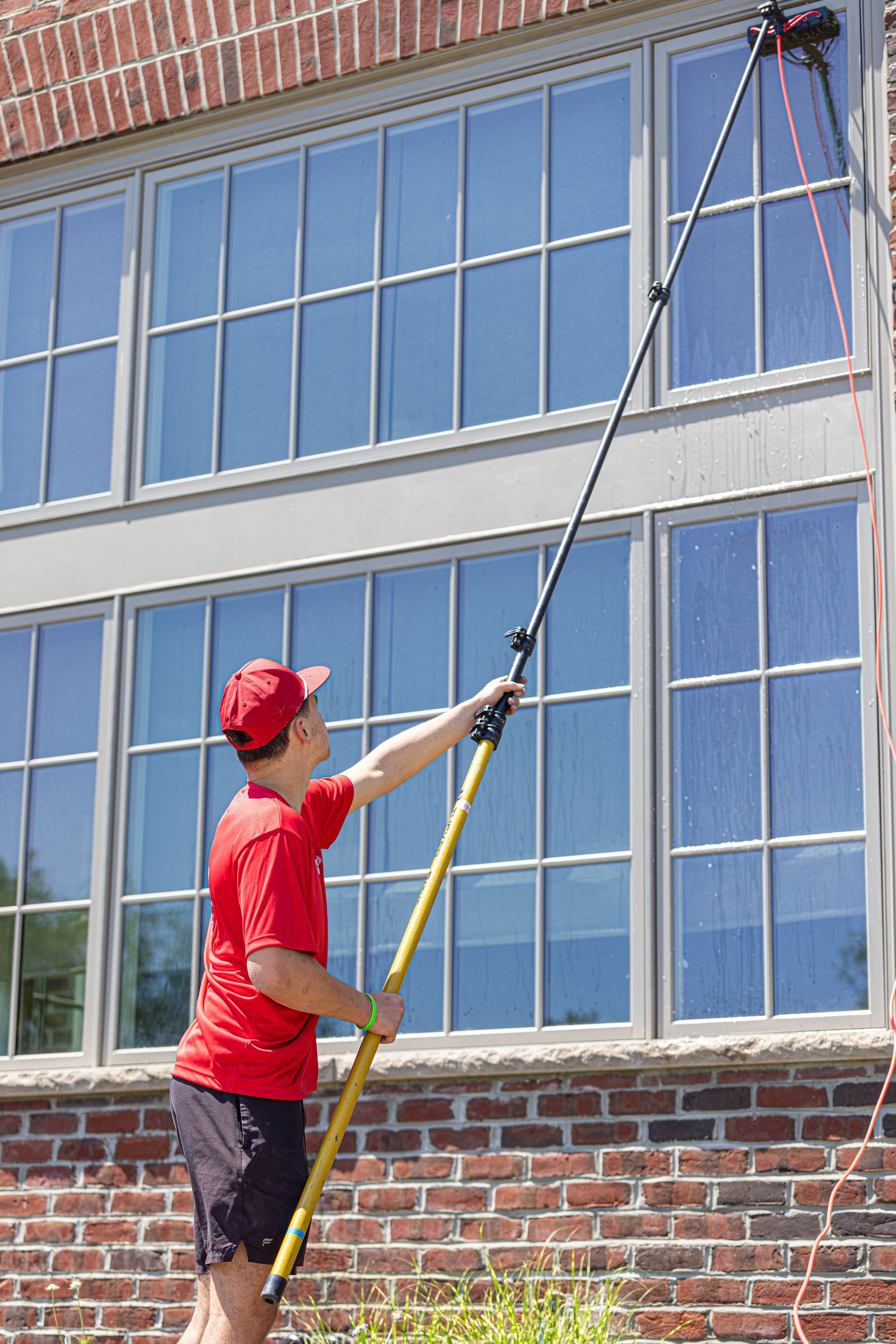 Worker in red shirt cleaning upper windows with a long pole beside a brick building