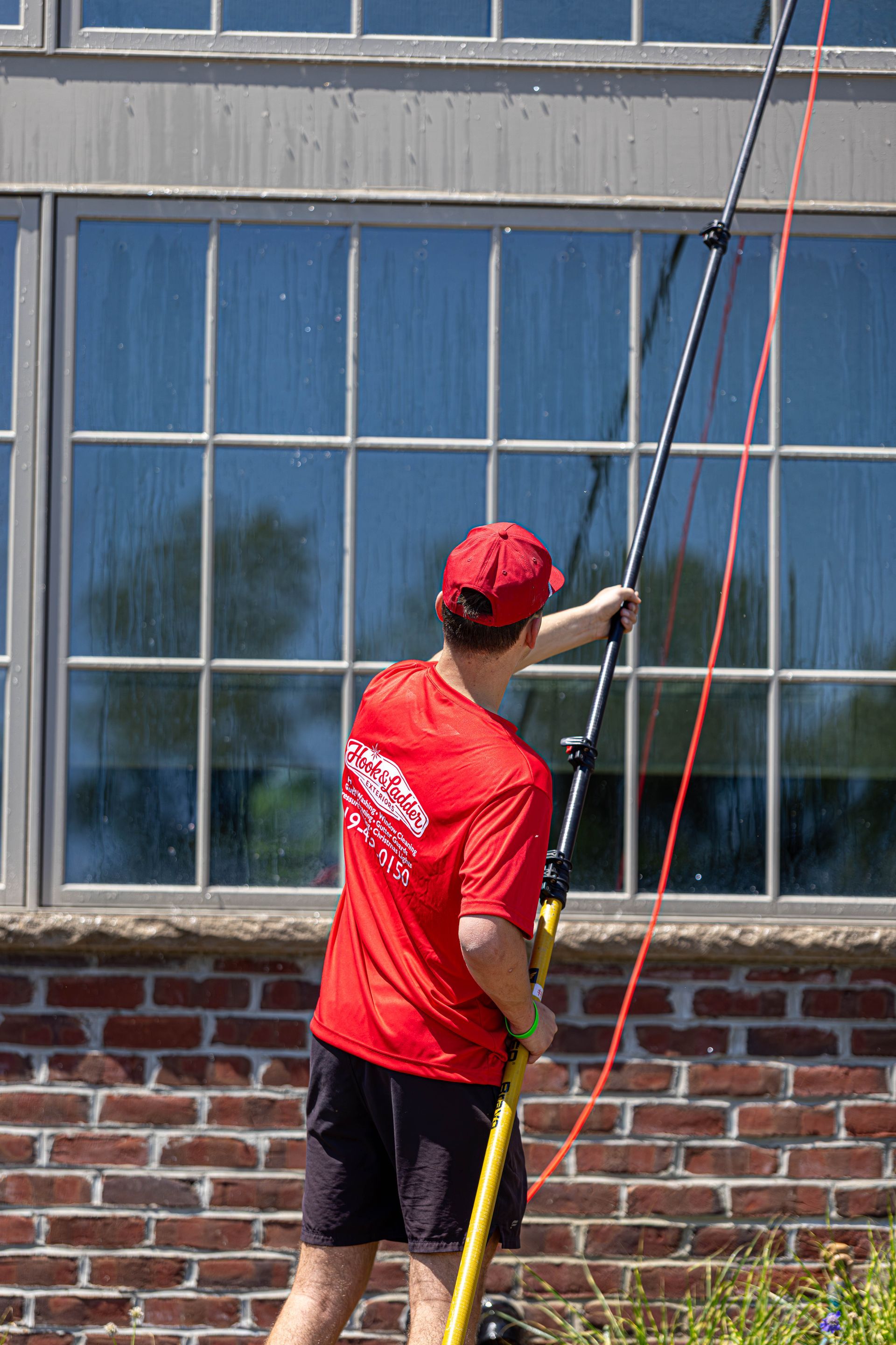 Person in a red shirt using a long pole to clean a large window on a brick building.
