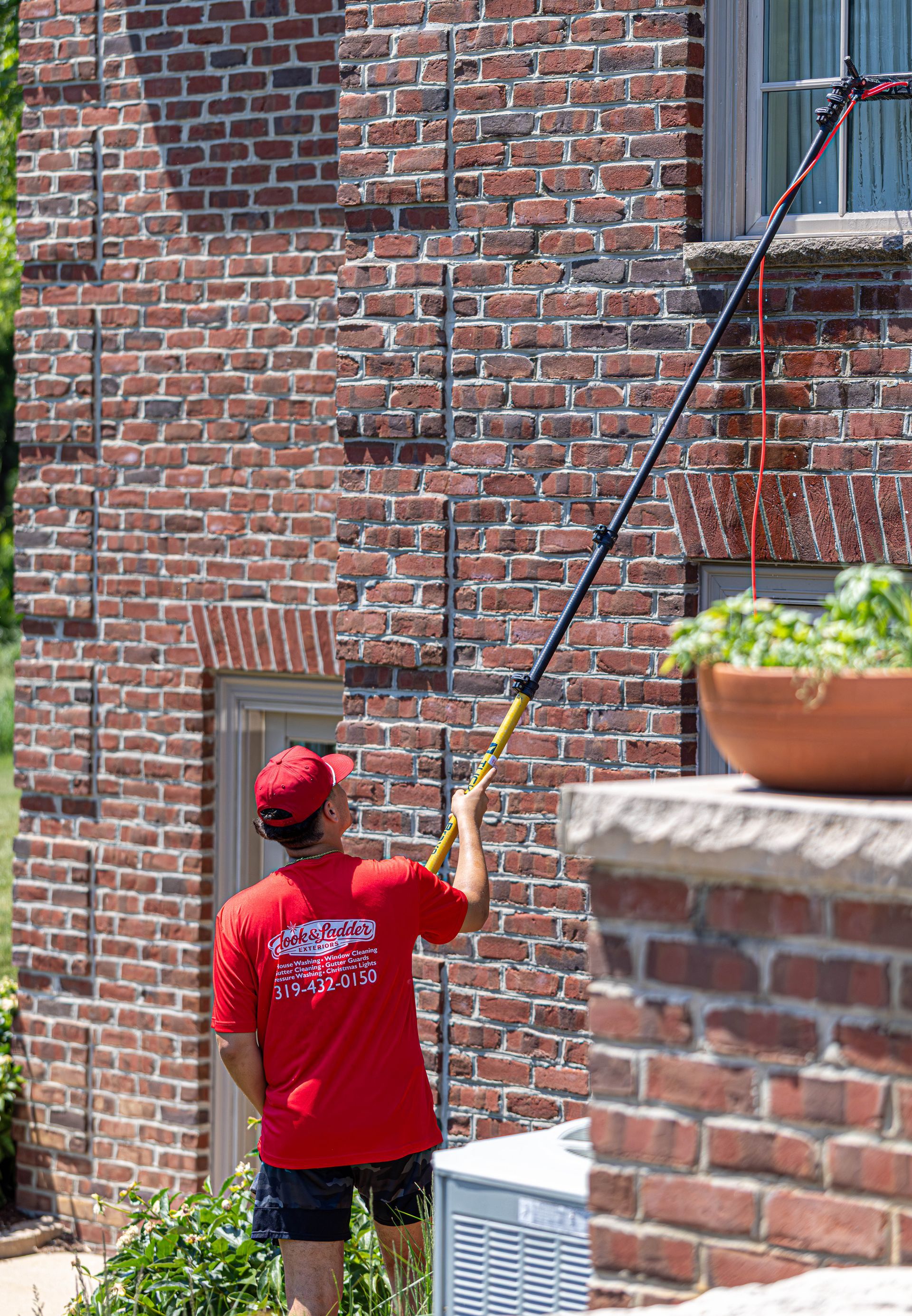 Worker in red shirt using a long pole near a brick building with a window planter.
