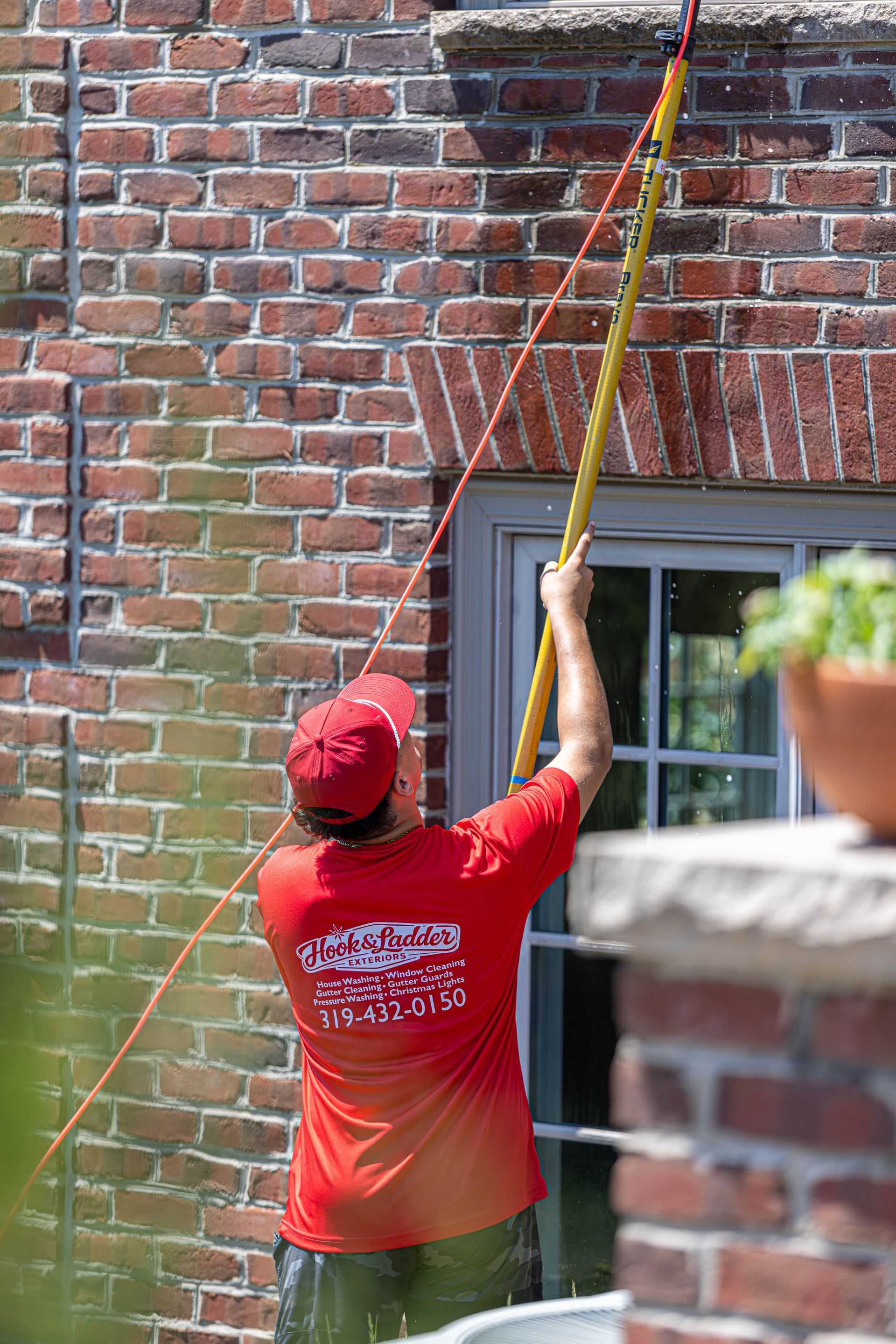 Worker in red shirt using a long pole to inspect or clean a brick wall above a window.