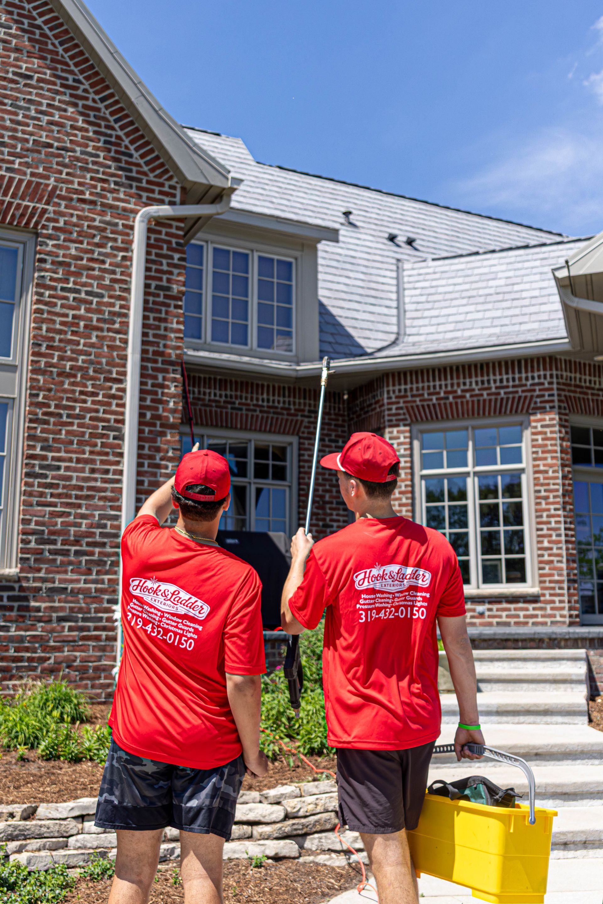 Two workers in red shirts carry a yellow tool case up steps to a brick house.
