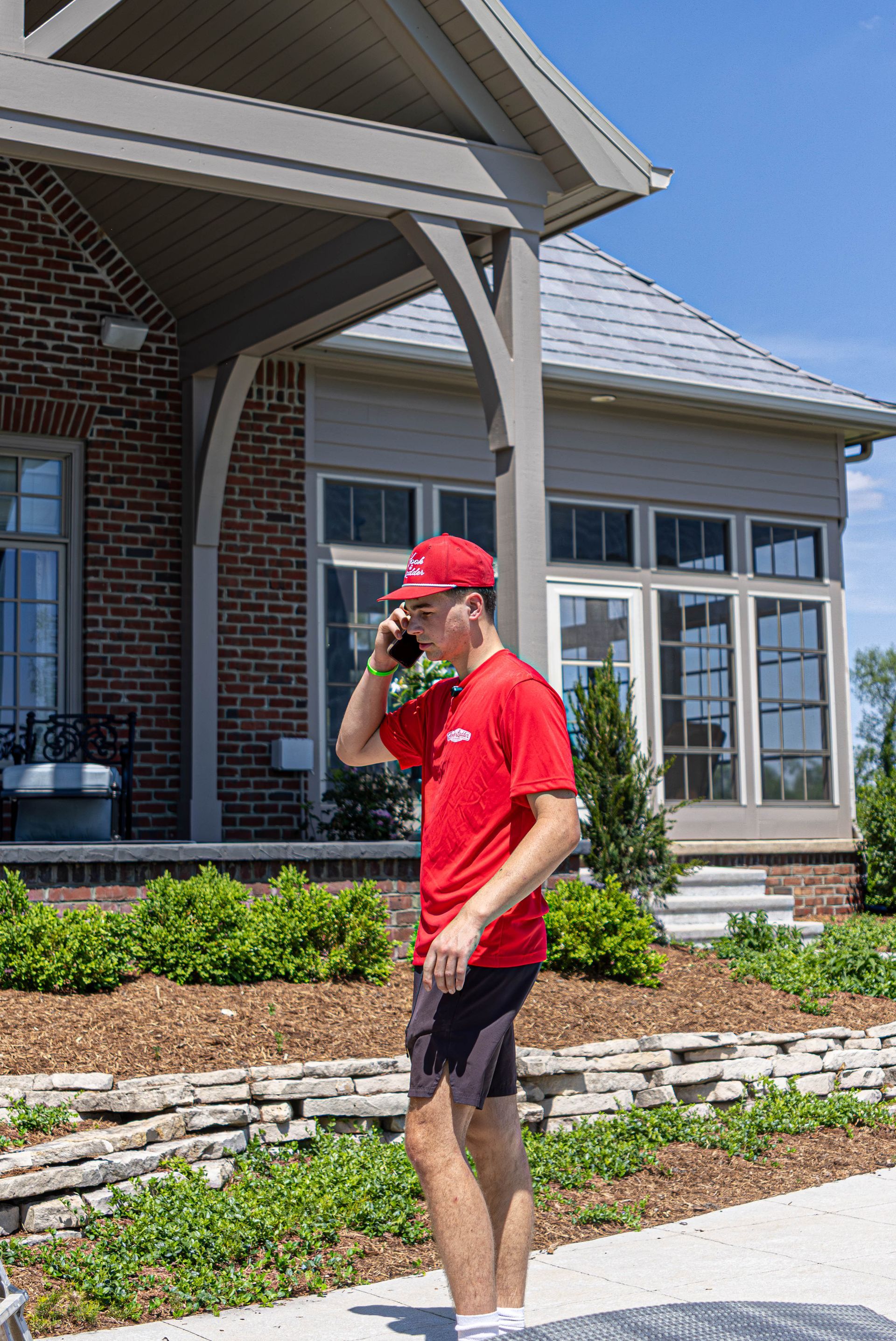 Person in red shirt and cap standing outside a brick house, using a phone on a sunny day