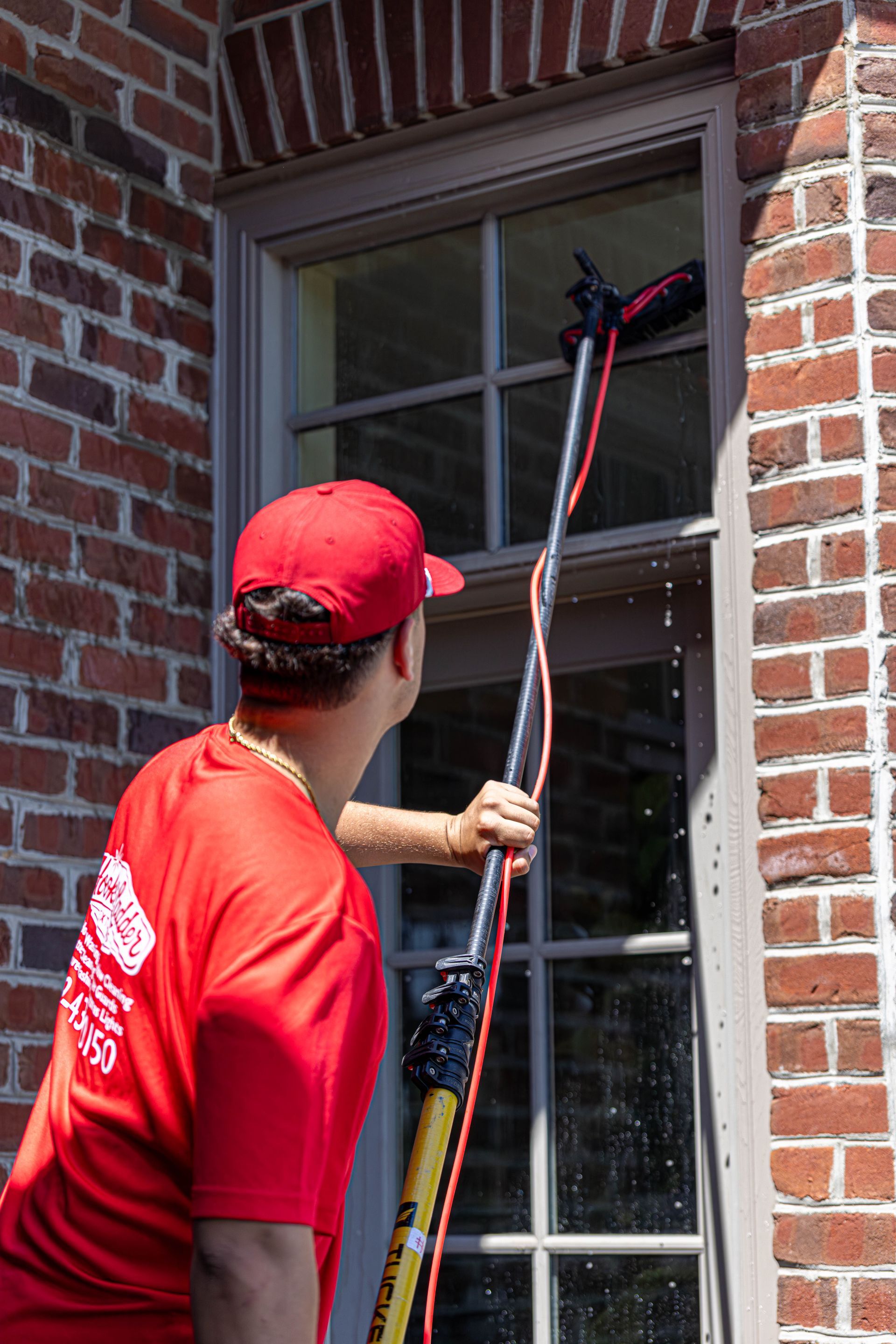 Person cleaning an upper window with a long-handled squeegee beside a brick wall