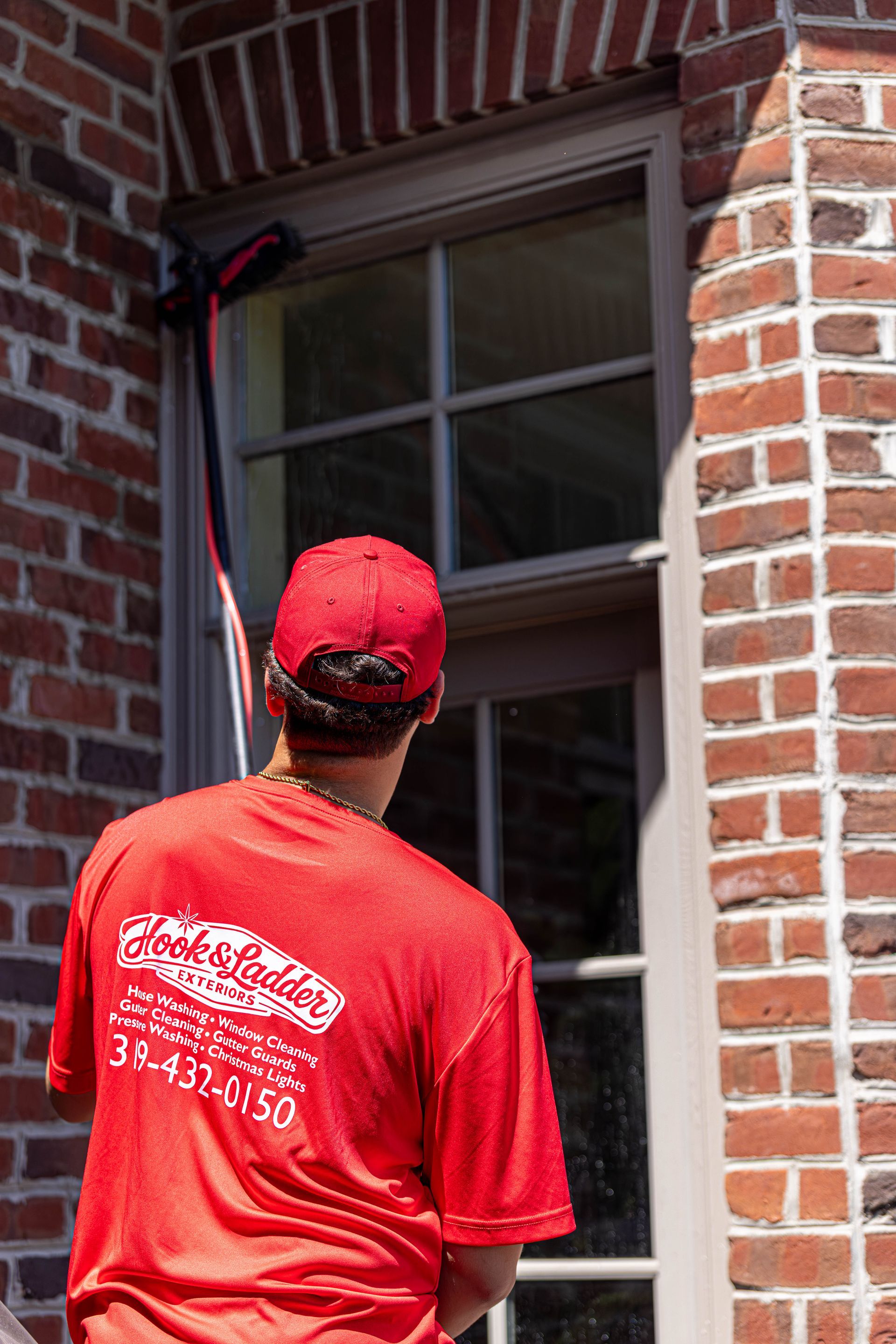 Worker in red shirt and cap cleaning an exterior window with a long pole.