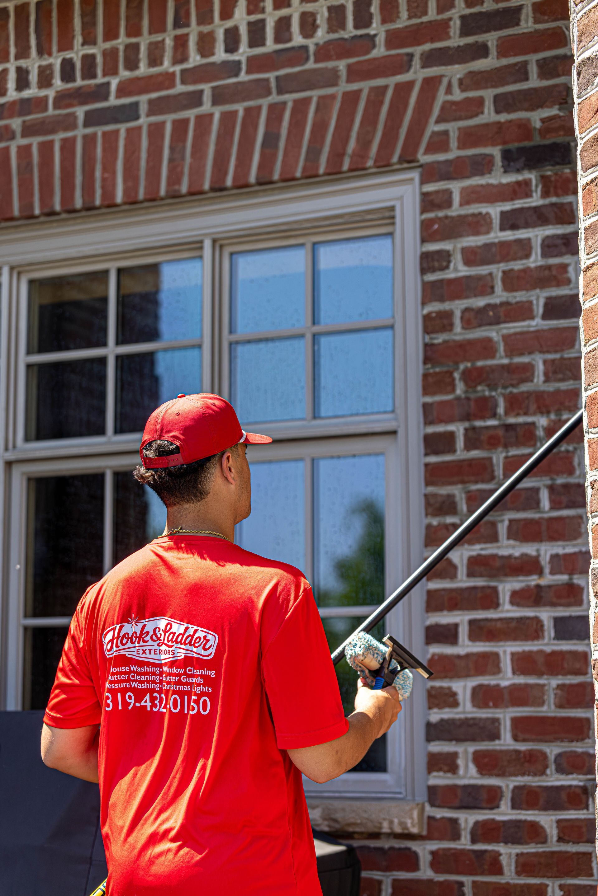 Person in a red shirt using a pressure washer on a brick house wall near a window.