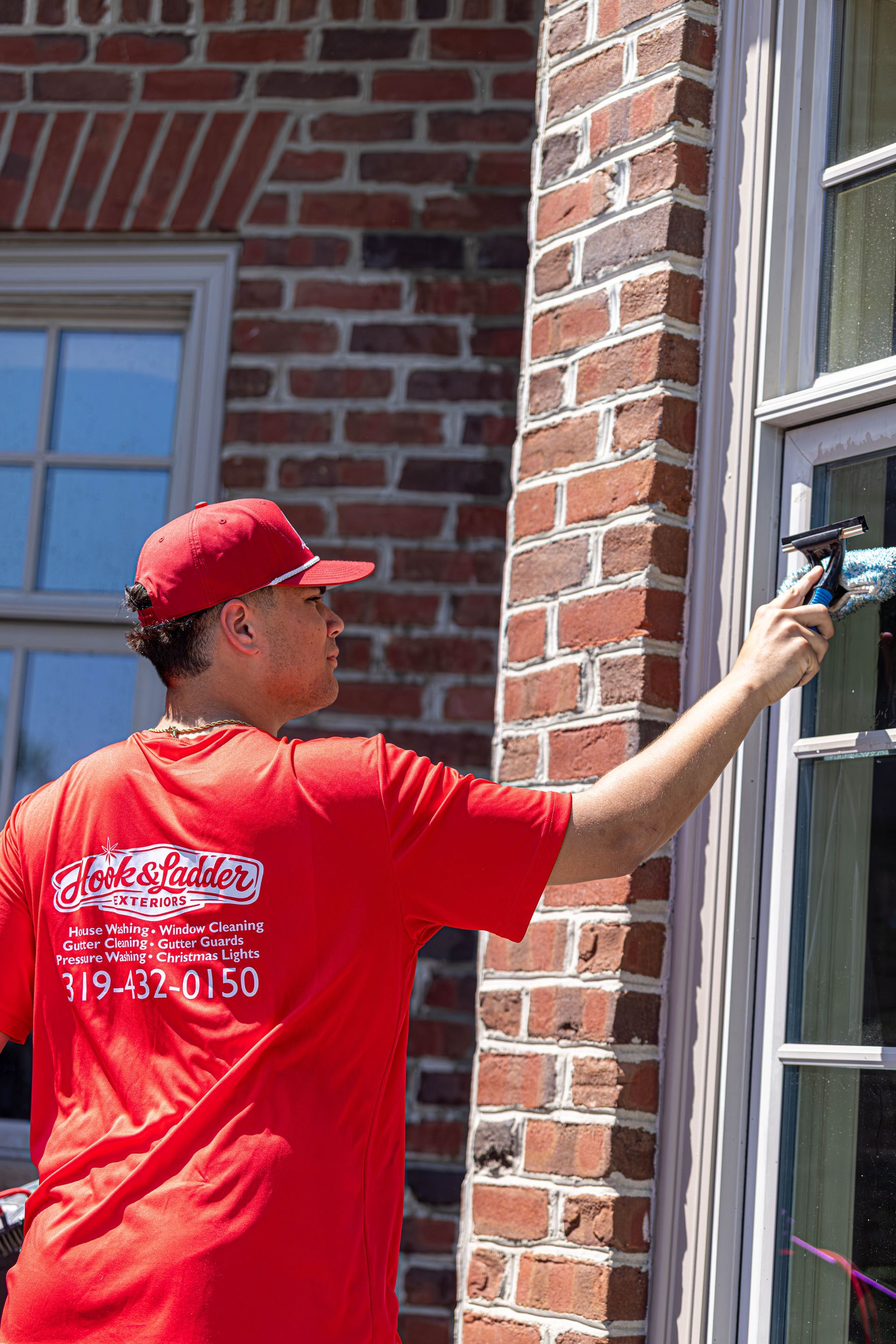 Person in a red shirt and cap applying caulk to a brick wall beside a window