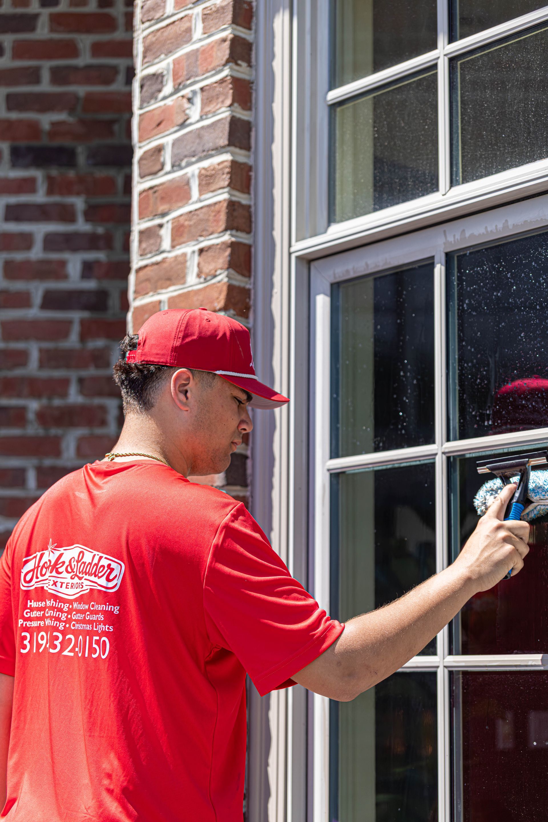 Person in red shirt and cap using a sprayer to clean a window on a brick building