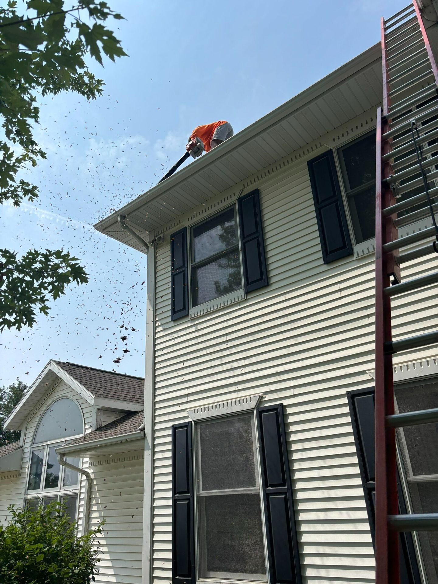 Worker clearing debris from a roof beside a house with a ladder attached.