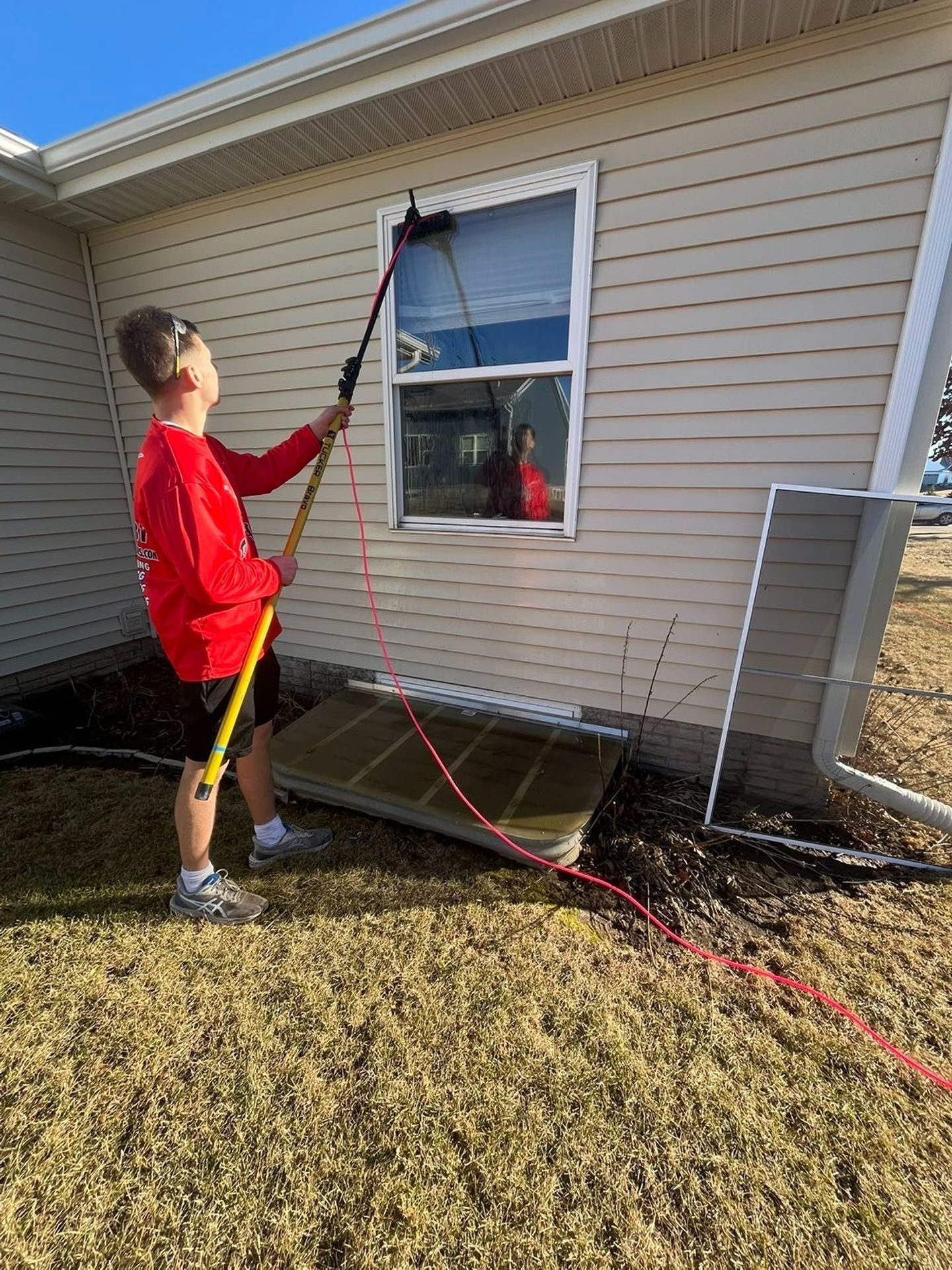 Person in a red shirt uses a long pole to clean an exterior house window.