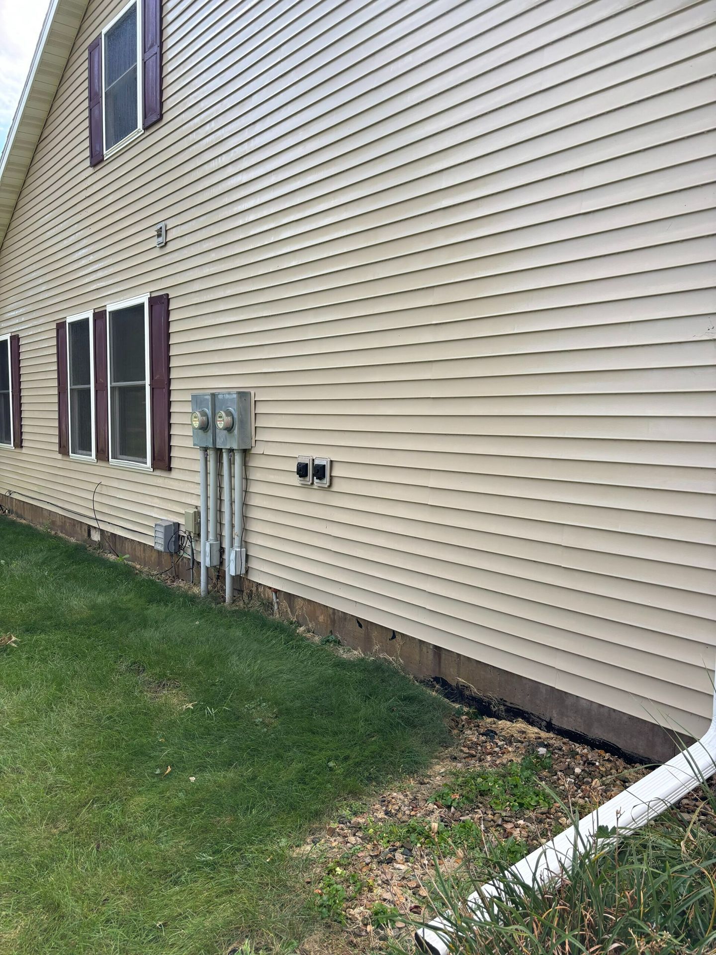 Side view of a beige house with windows, utility meters, and a white downspout beside a grassy yard