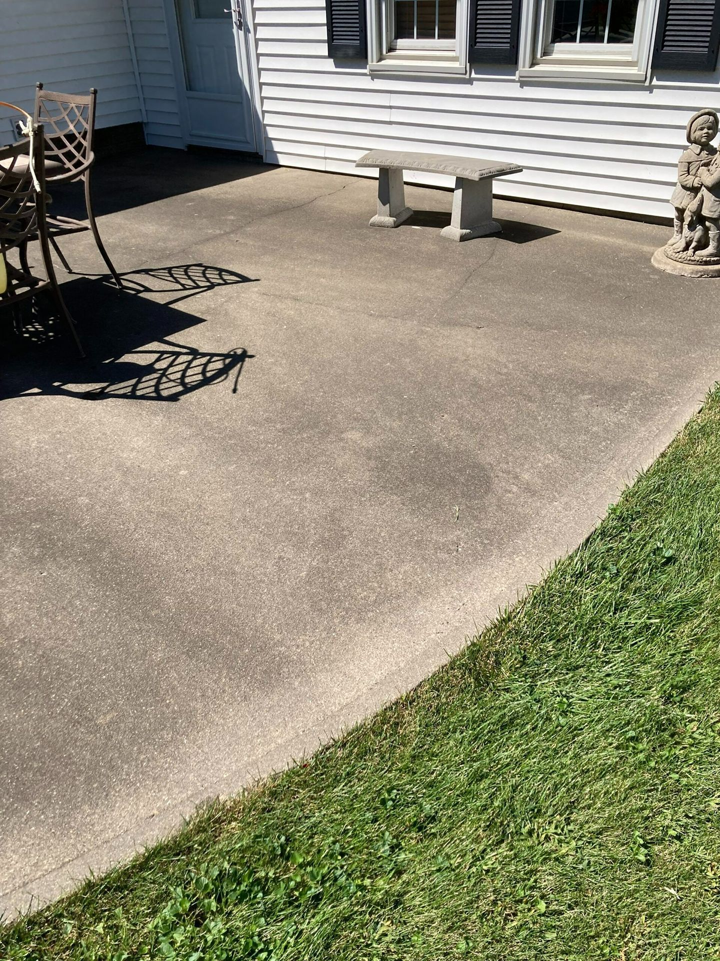 Concrete patio beside a house, with a bench, outdoor table and chairs, and a strip of grass.