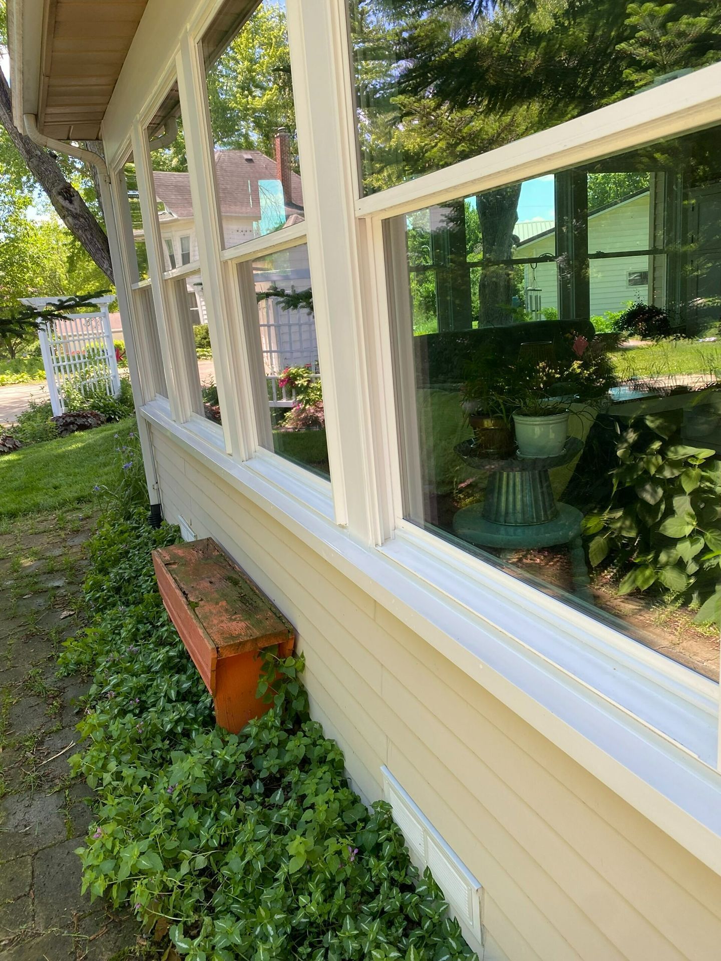 Side of cream house with white-trimmed windows, reflecting greenery and a brick planter below.