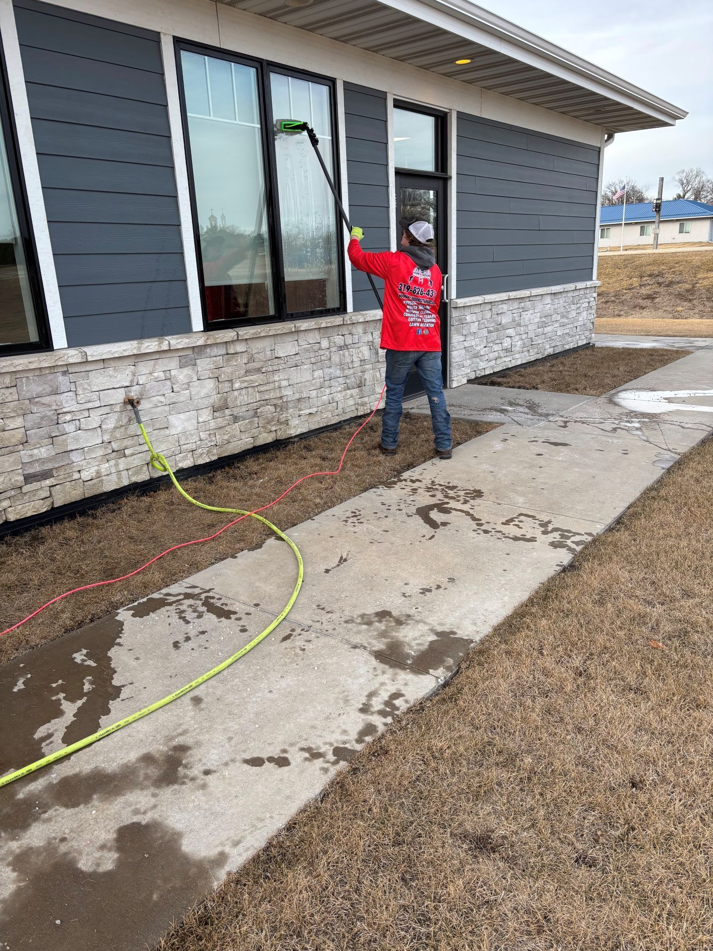 Worker in red shirt spraying water beside a modern house with a wet concrete walkway.