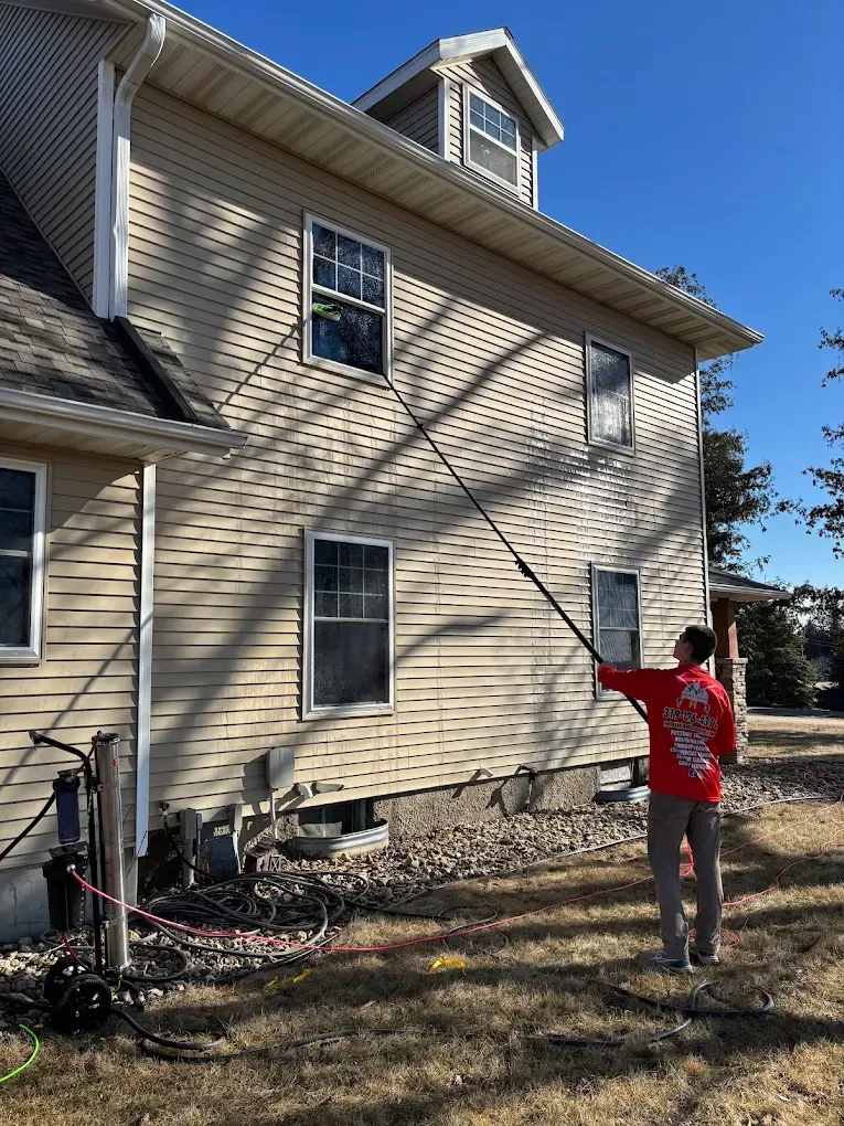 Person in red using a long pole to clean a second-story house window under a clear blue sky