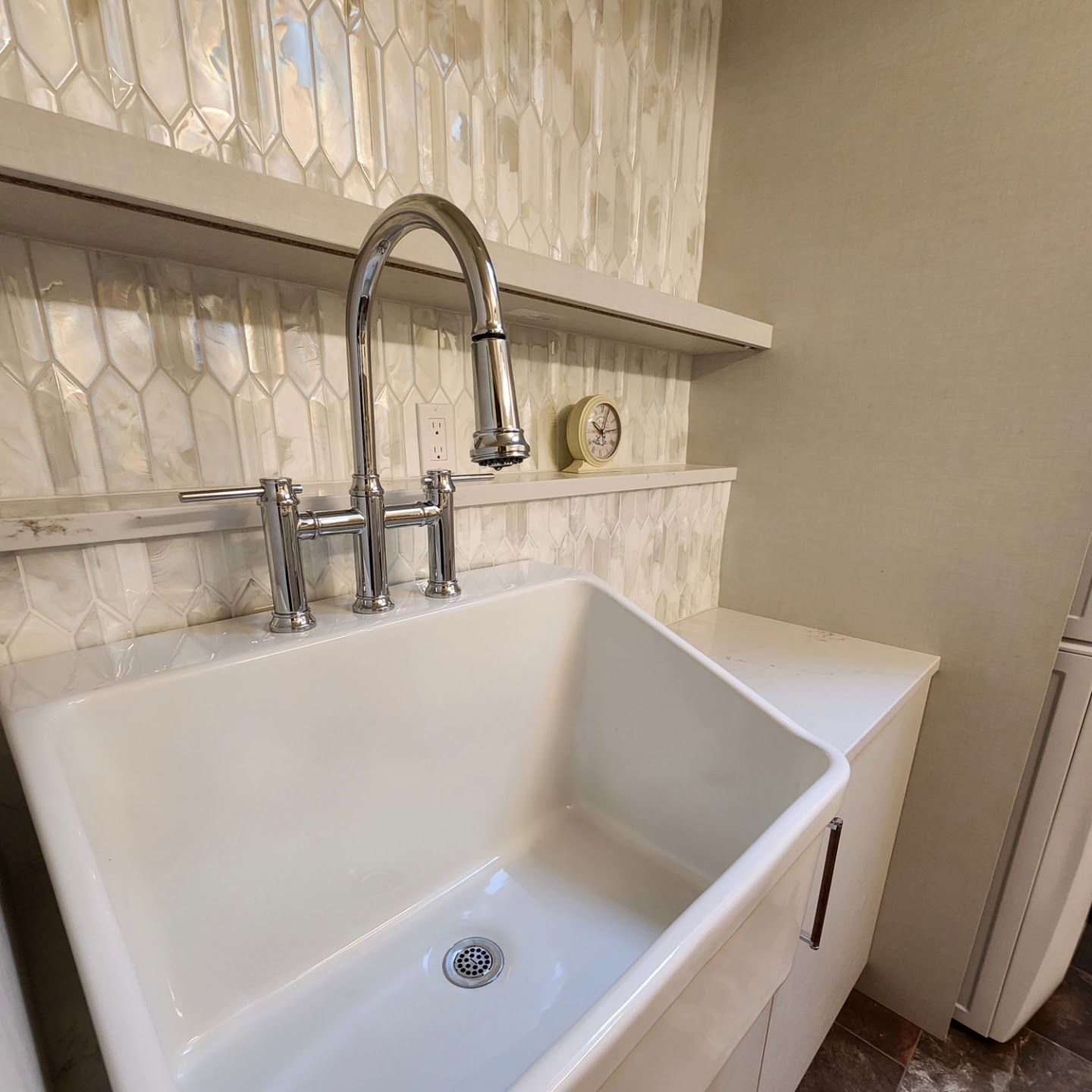 White farmhouse sink with chrome faucet, set against a tiled backsplash and floating shelf in a kitchen.